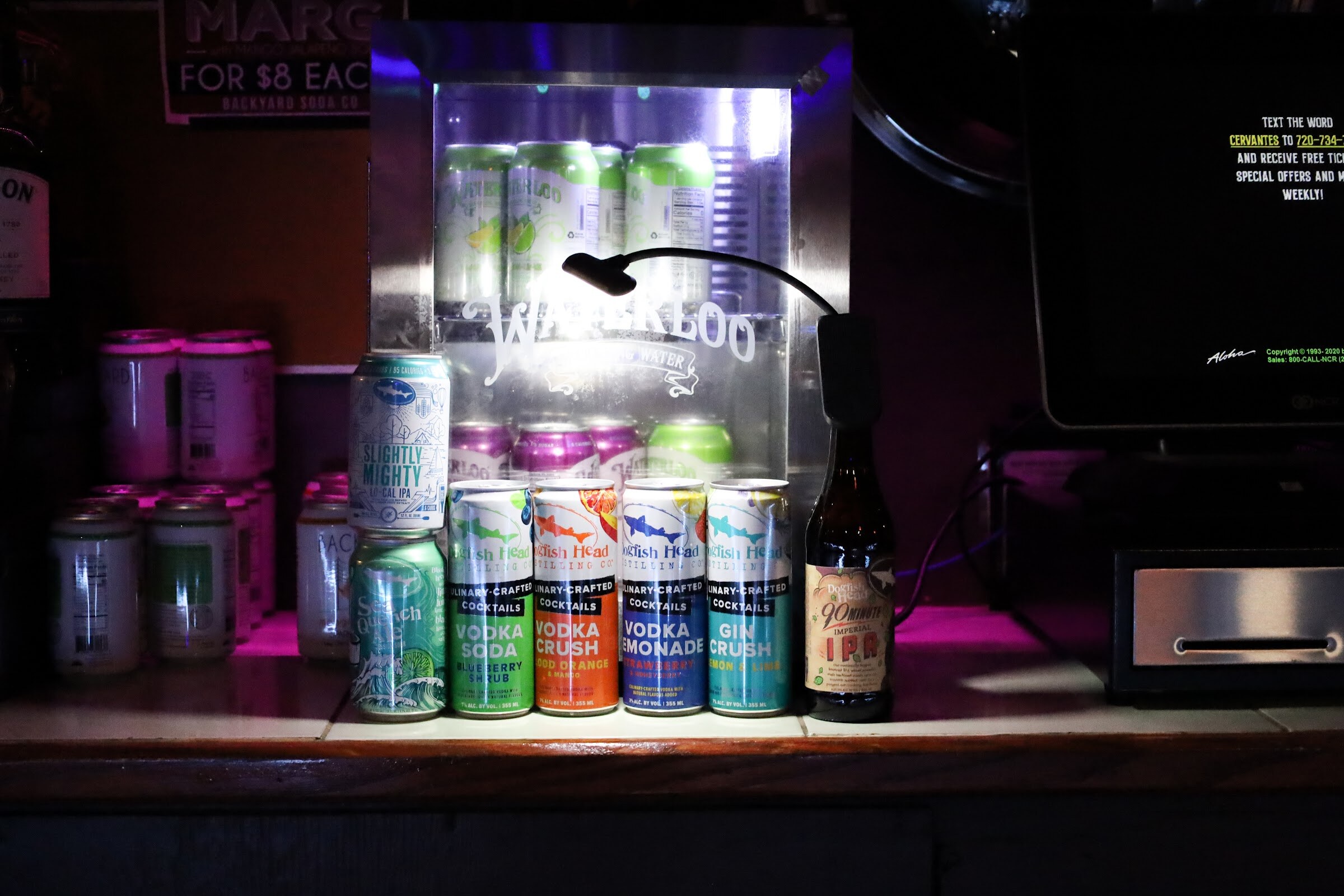 Cans of Dogfish Head Vodka Soda, Vodka Crush, Vodka Lemonade and Gin Crush and a bottle of 90 Minute Imperial IPA stand in front of a lit Waterloo sparkling water cooler beside a can of Slightly Mighty.