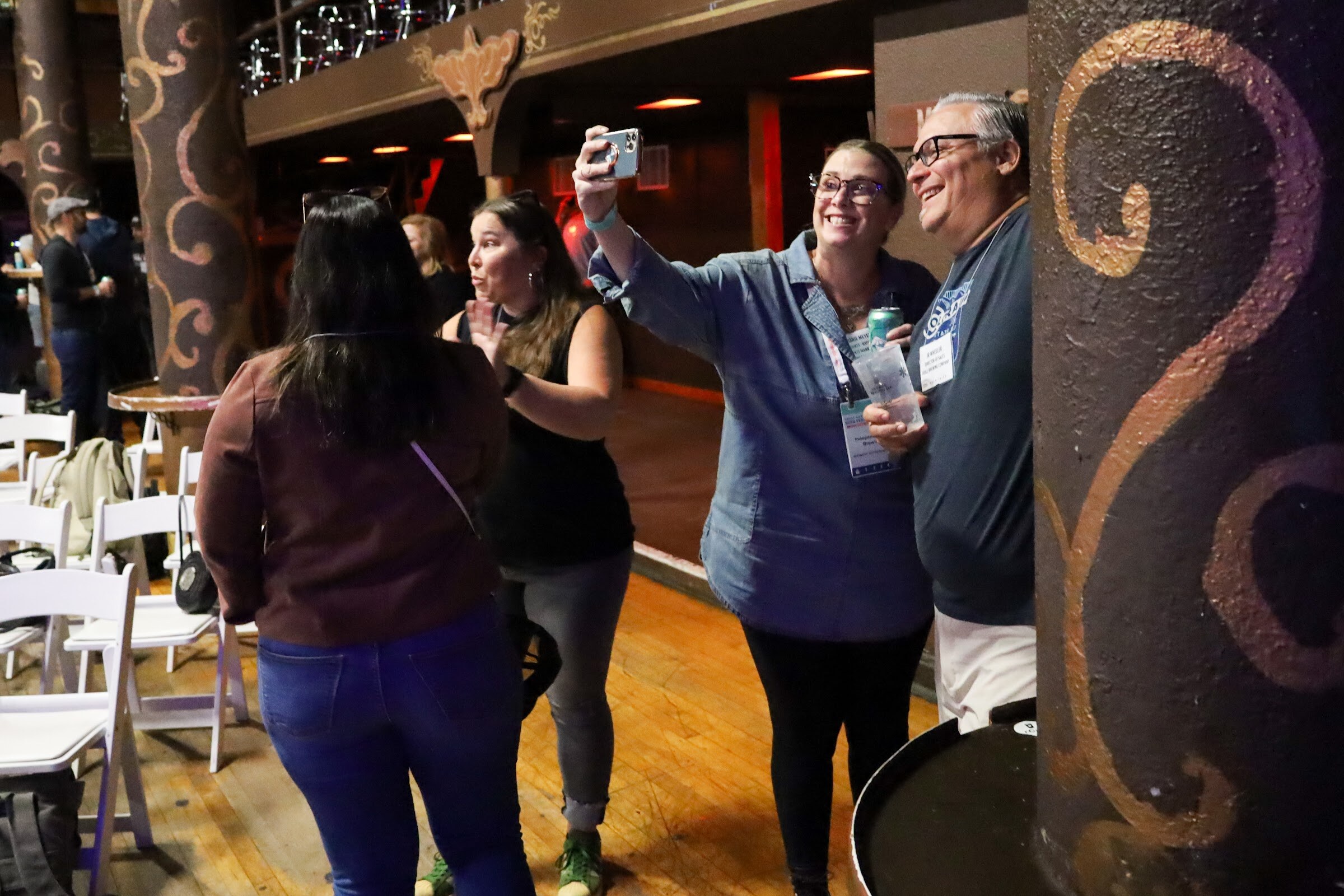 A woman in a denim shirt holds up a phone to take a selfie with a smiling man in glasses beside an ornate painted pillar, while a woman in a brown jacket and a woman in black talk to the left.
