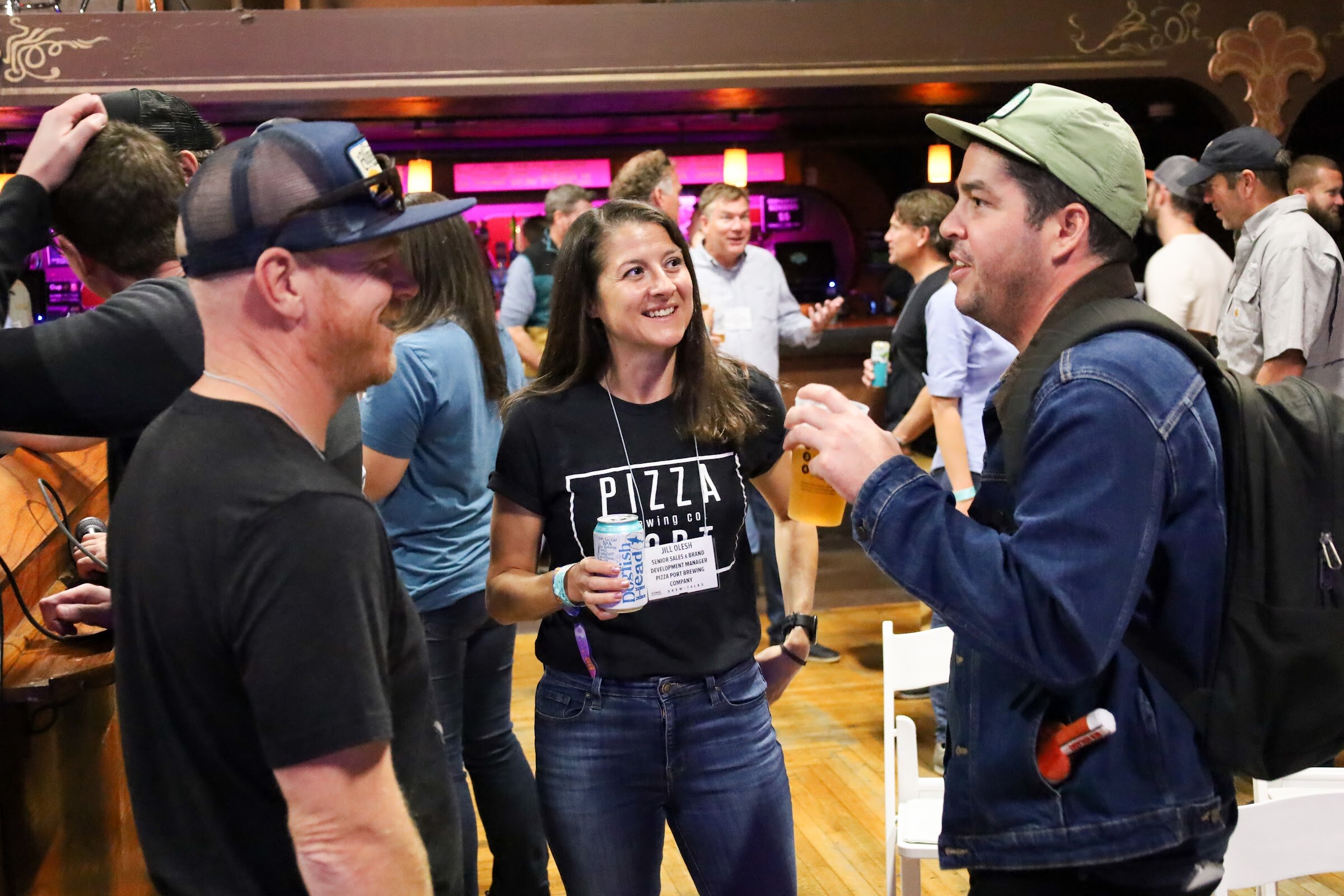 A smiling woman in a black Pizza Brewing Co. t-shirt holding a can talks with a man in a green cap and denim jacket with a backpack who holds a beer, while a man in a blue trucker cap and a black t-shirt stands in the foreground.