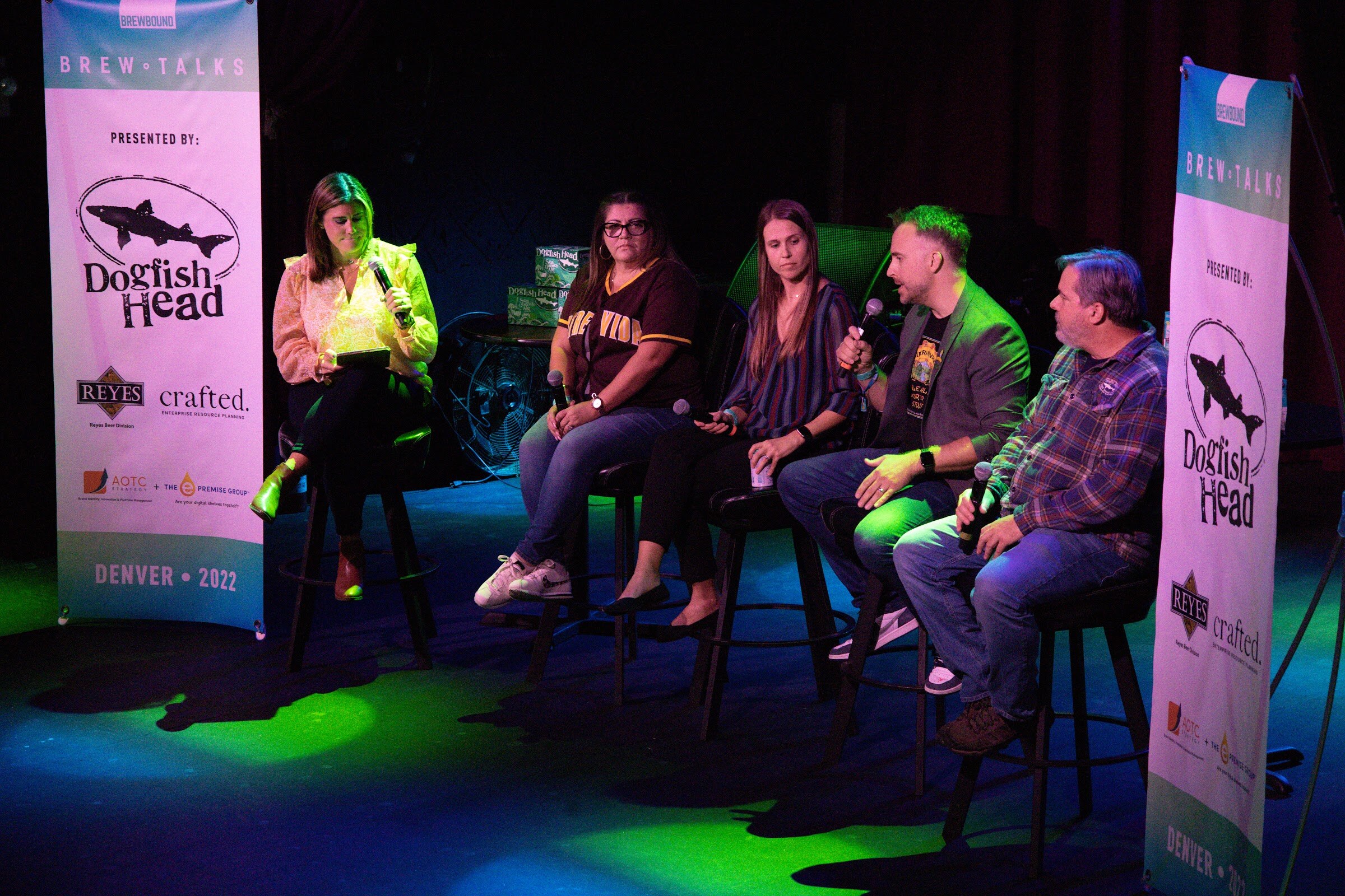A woman in a peach ruffled blouse holds a tablet and microphone beside a woman in a brown baseball jersey and glasses, a woman in a striped blouse, a man with green-tinted hair in a gray blazer speaking, and a man in a plaid shirt, on bar stools beside a Dogfish Head banner.
