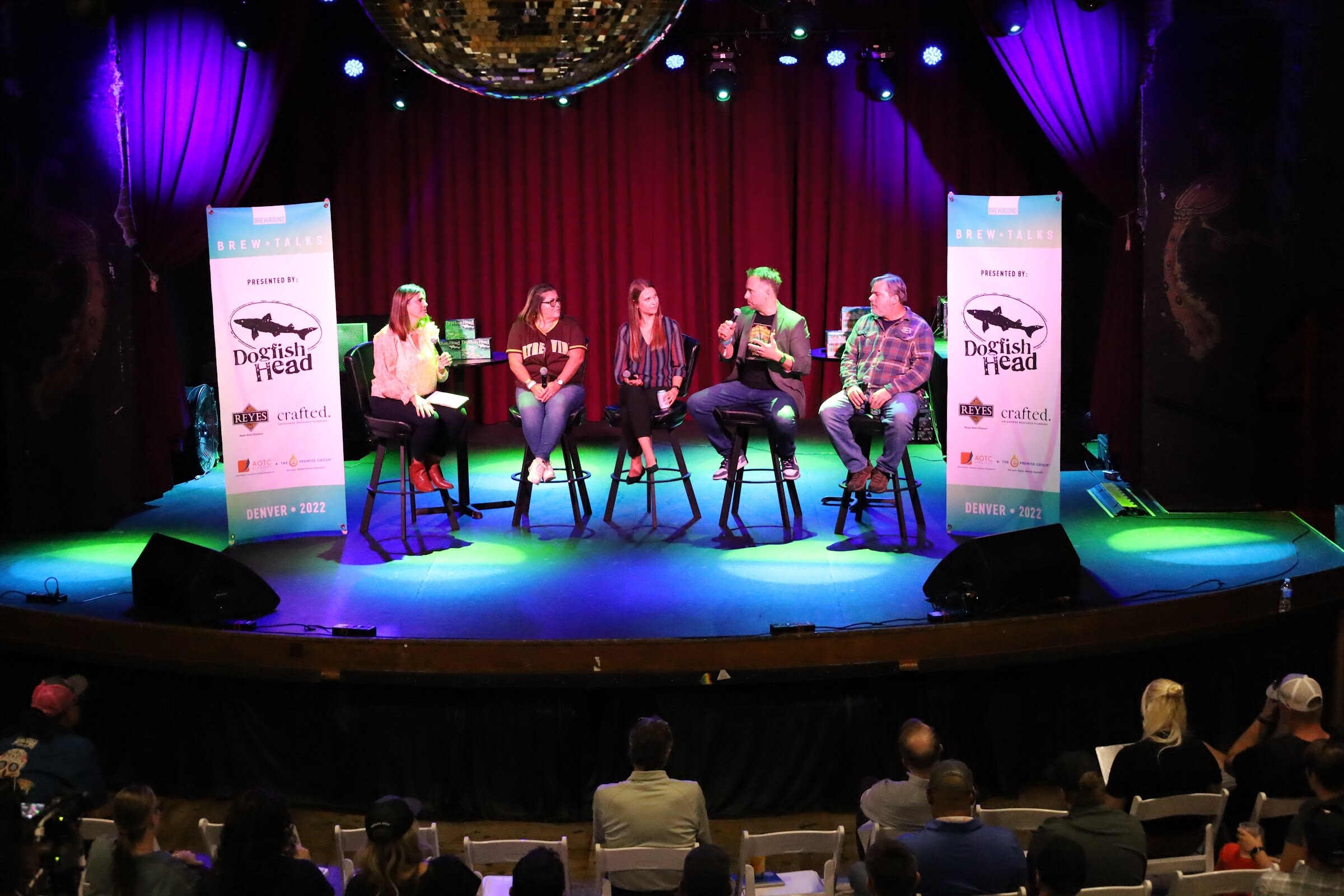 Wide view of a stage lit in blue and green with five people on black bar stools between two Brew Talks banners presented by Dogfish Head, a disco ball at the top of the frame and the backs of audience members at the bottom.