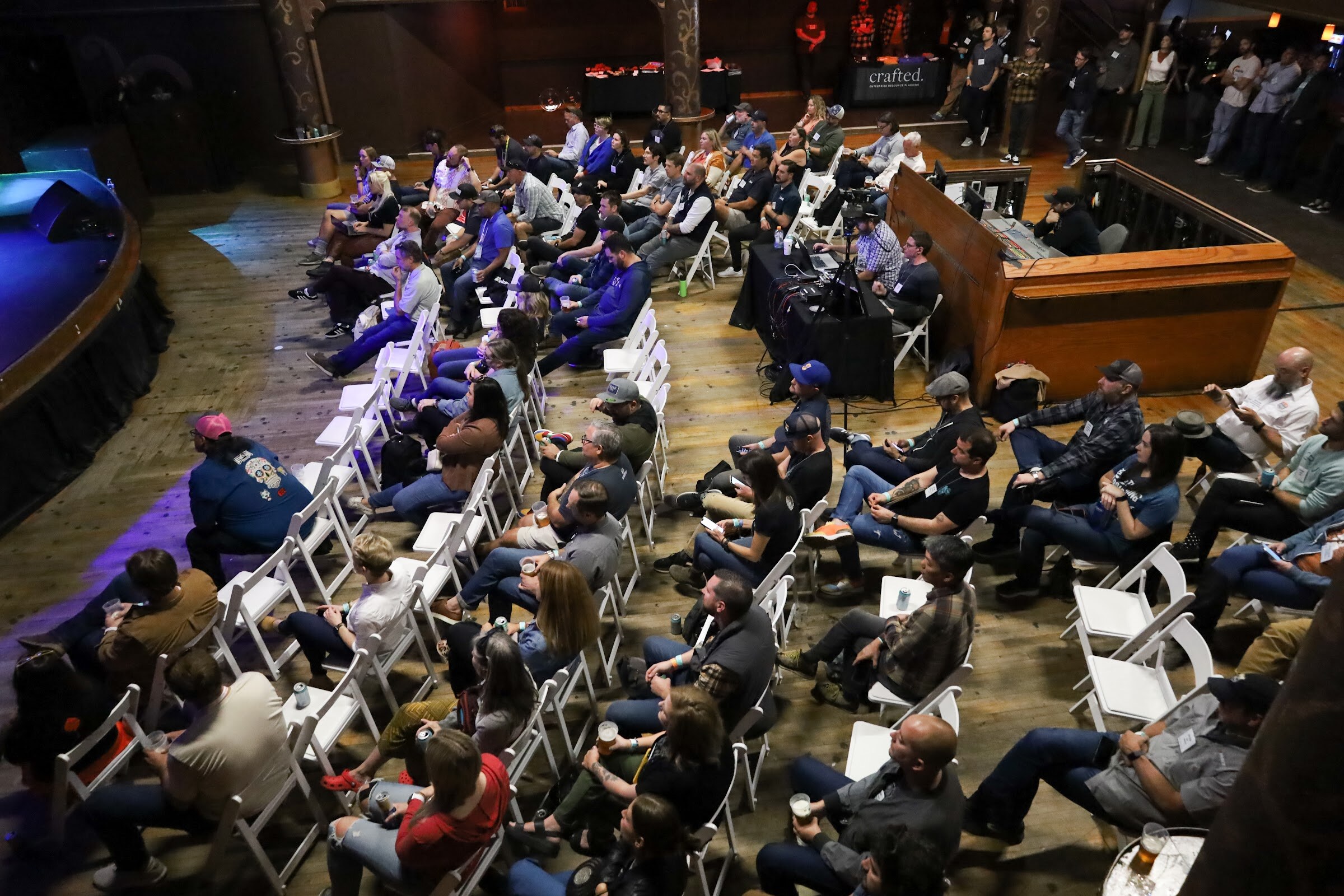 High-angle view of seated attendees with name badges in white folding chairs, with a sound desk and standing guests at the back and the edge of the stage at the left.