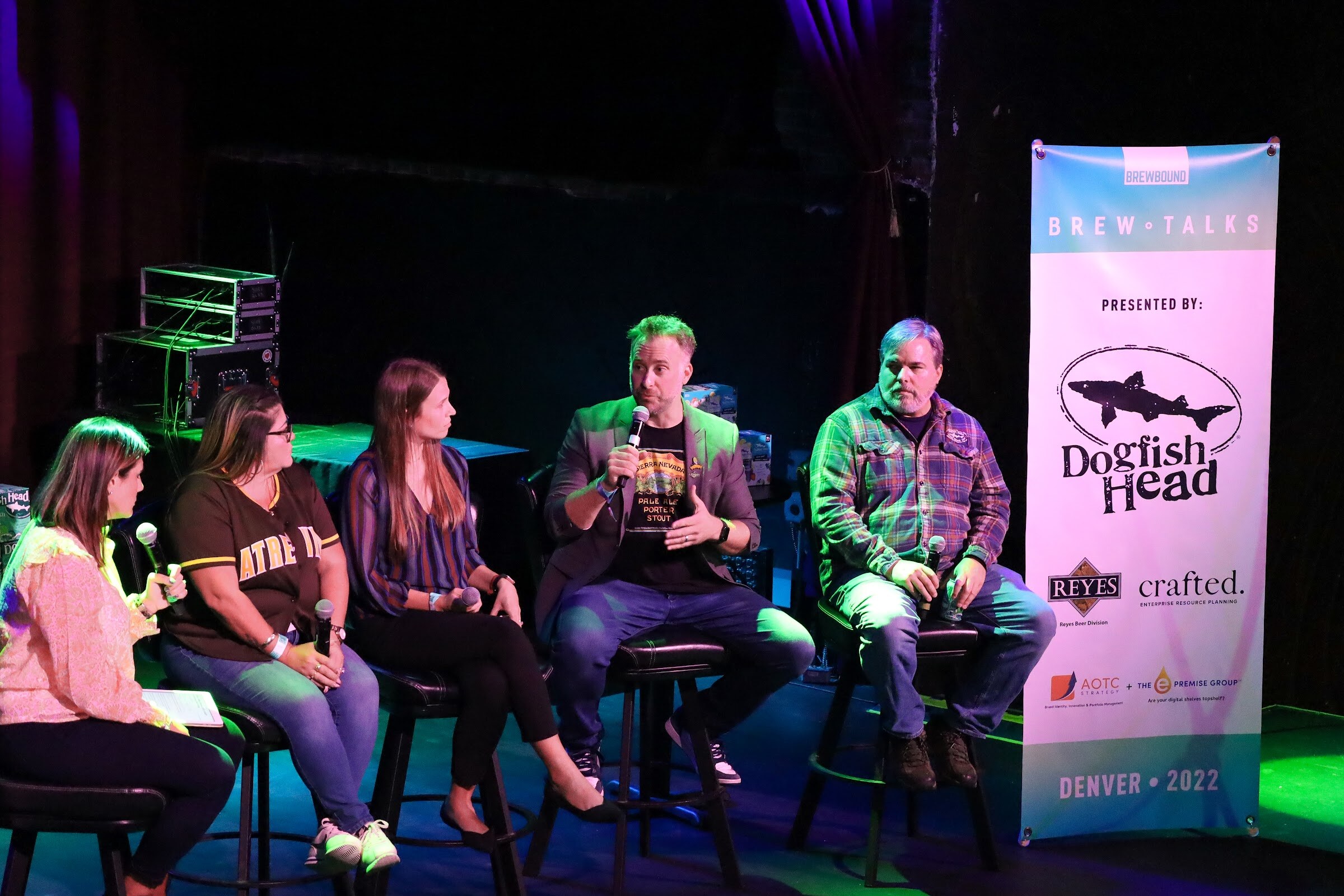 A man with green-tinted hair in a gray blazer and a graphic t-shirt speaks into a microphone, with a woman in a peach blouse, a woman in a brown jersey, a woman in a striped blouse and a man in a plaid shirt seated beside him, next to a Brew Talks banner.