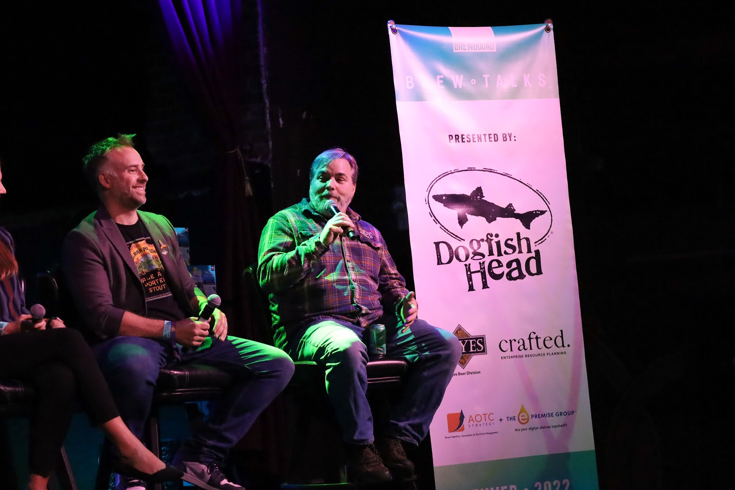 A man in a green plaid shirt speaks into a microphone while a smiling man with green-tinted hair in a gray blazer and graphic t-shirt sits beside him, next to a Brew Talks banner presented by Dogfish Head, Reyes and crafted.