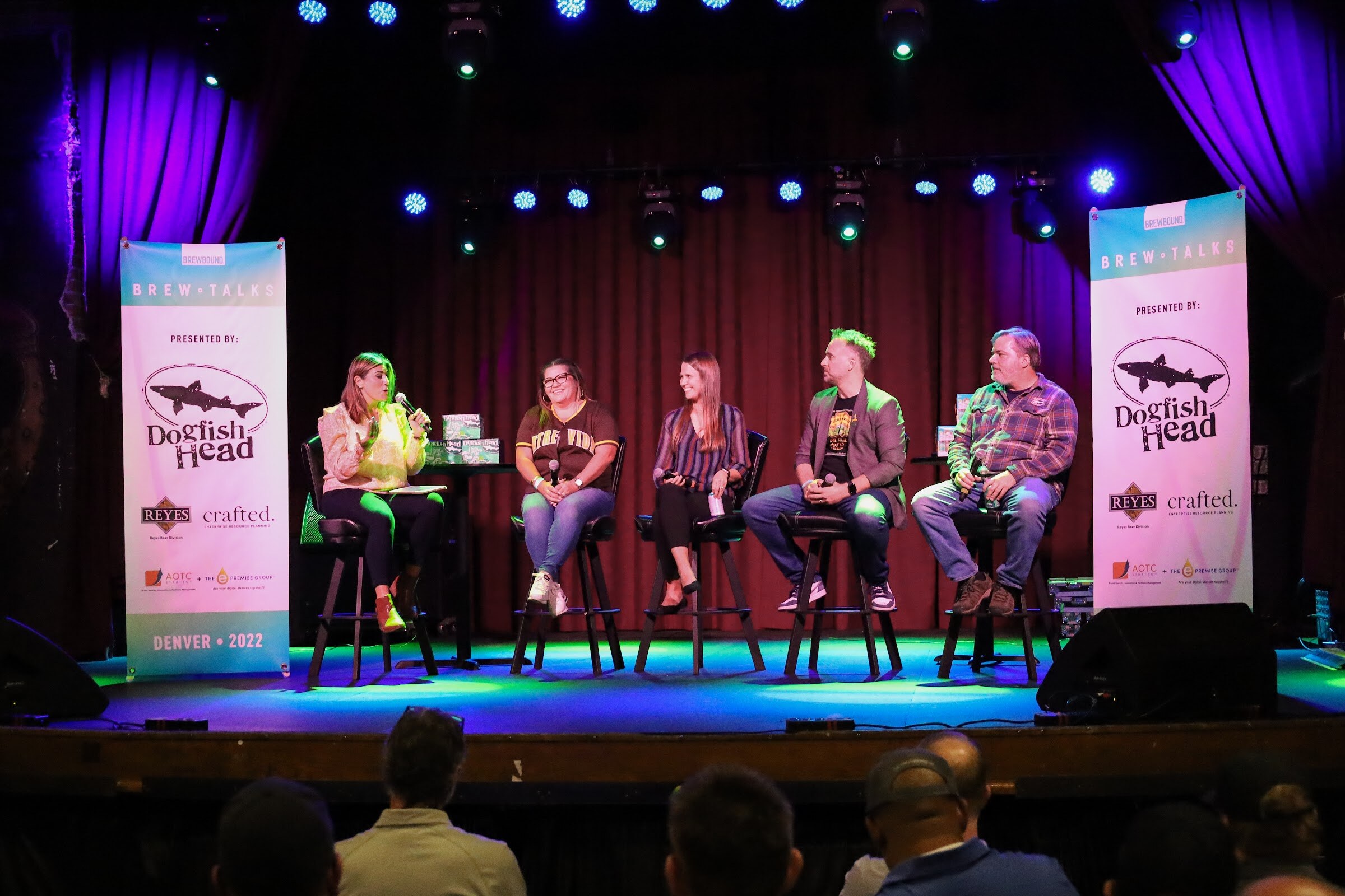 Five people on black bar stools on a stage lit in blue and green between two Brew Talks banners, with the backs of audience members' heads along the bottom edge.