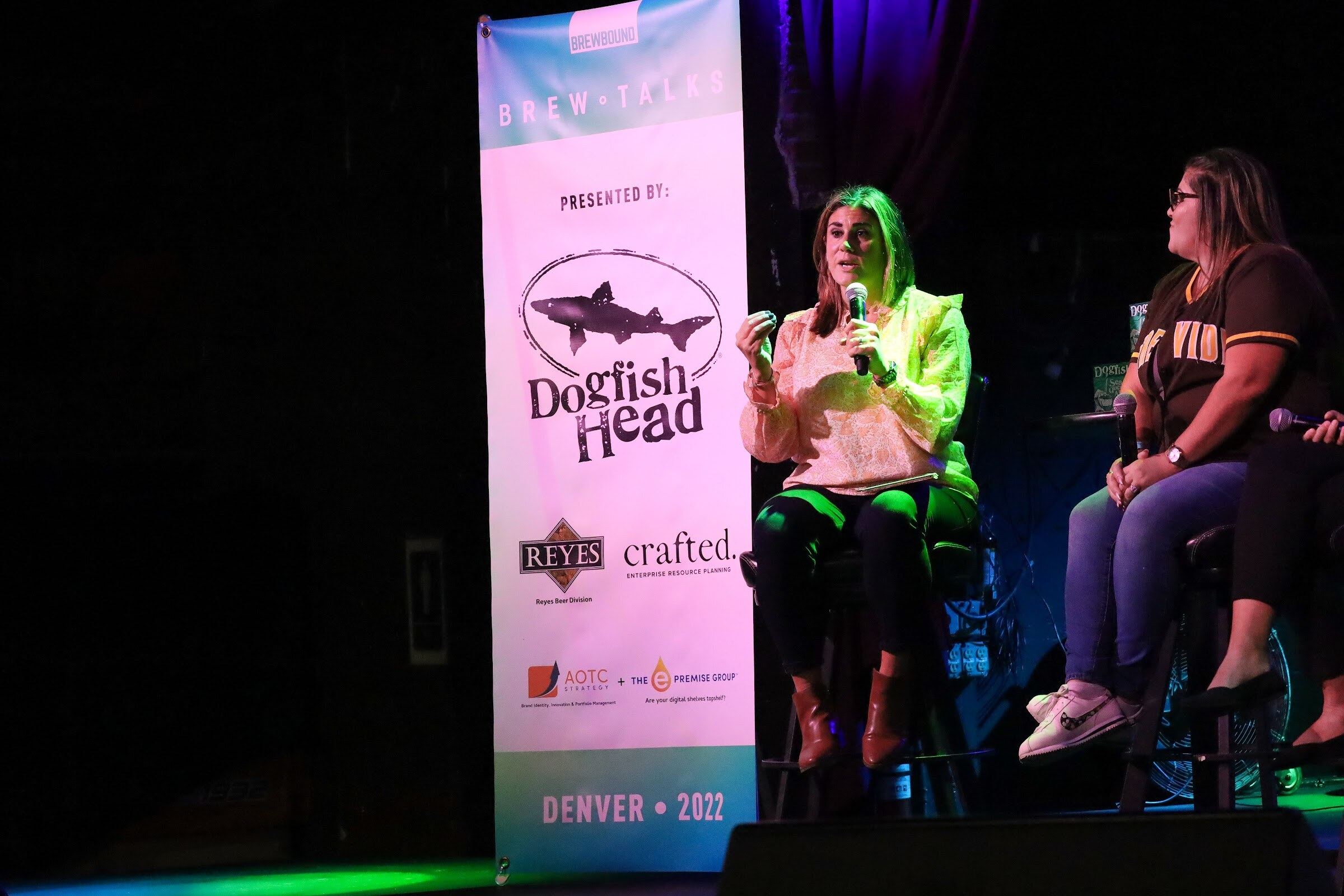 A woman in a peach ruffled blouse and brown boots speaks into a microphone beside a Brew Talks banner, with a woman in a brown baseball jersey and glasses seated at the right edge of the frame.