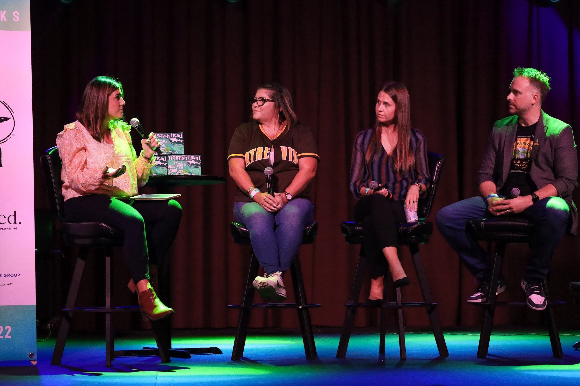 A woman in a peach ruffled blouse gestures with a microphone toward a woman in a brown jersey and glasses, a woman in a striped blouse and a man with green-tinted hair in a gray blazer, seated on bar stools in front of a dark curtain.