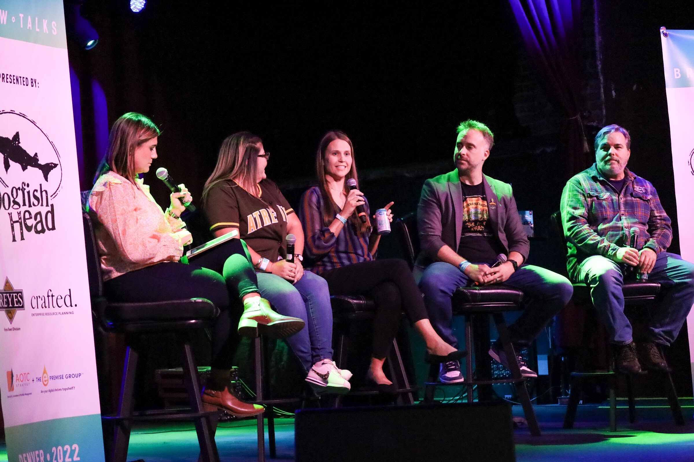 A woman in a striped blouse speaks into a microphone, with a moderator in a peach blouse, a woman in a brown jersey, a man in a gray blazer and a man in a green plaid shirt seated beside her next to a Brew Talks banner.