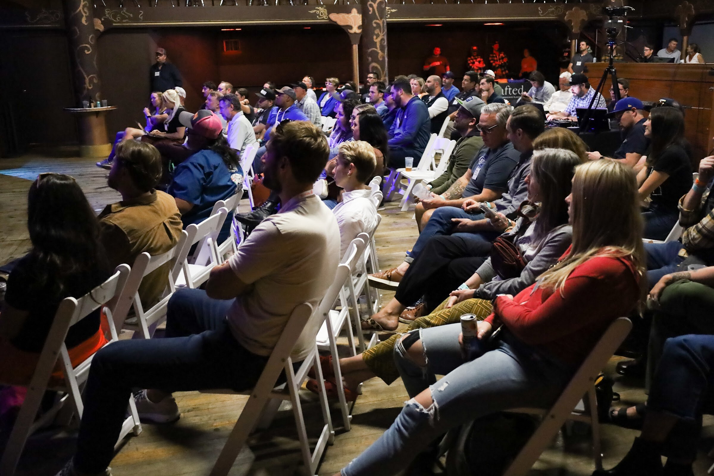 Seated attendees in white folding chairs, including a woman in a red top holding a can and a man in a cream t-shirt, face the stage in a dim room with a camera on a tripod and the bar behind them.