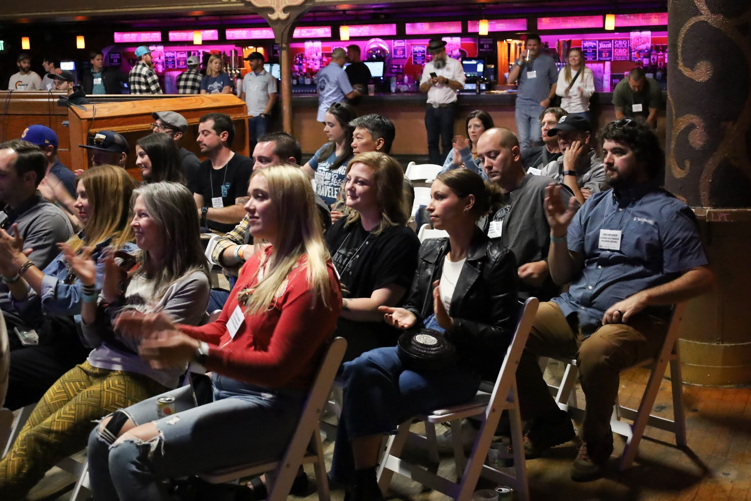 Seated attendees clap, including a woman in a red top, a woman in a black leather jacket and a bearded man in a blue button-up shirt, with the pink-lit bar and standing guests behind them.