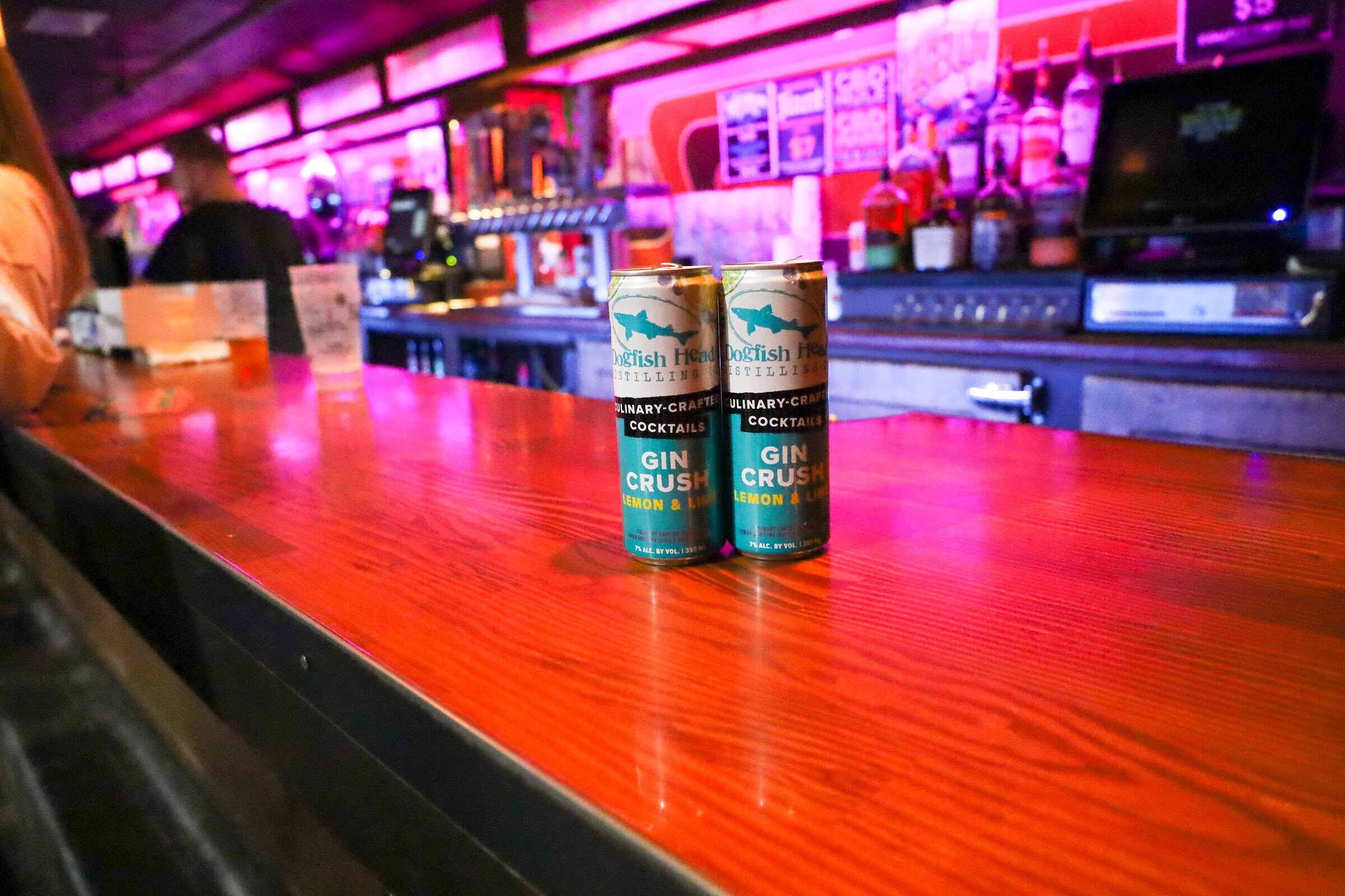 Two cans of Dogfish Head Distilling Culinary-Crafted Cocktails Gin Crush Lemon and Lime stand on a red-orange wooden bar, with bottles, taps and a register in the pink-lit background.