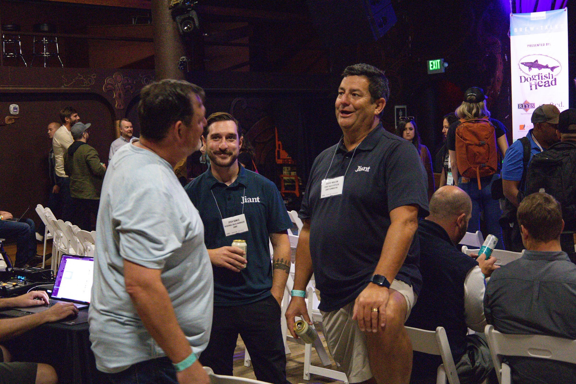 A man in a light gray t-shirt faces two men in navy Jiant polo shirts with name badges and cans in hand, with white folding chairs, a laptop at a sound desk and a Dogfish Head banner behind them.