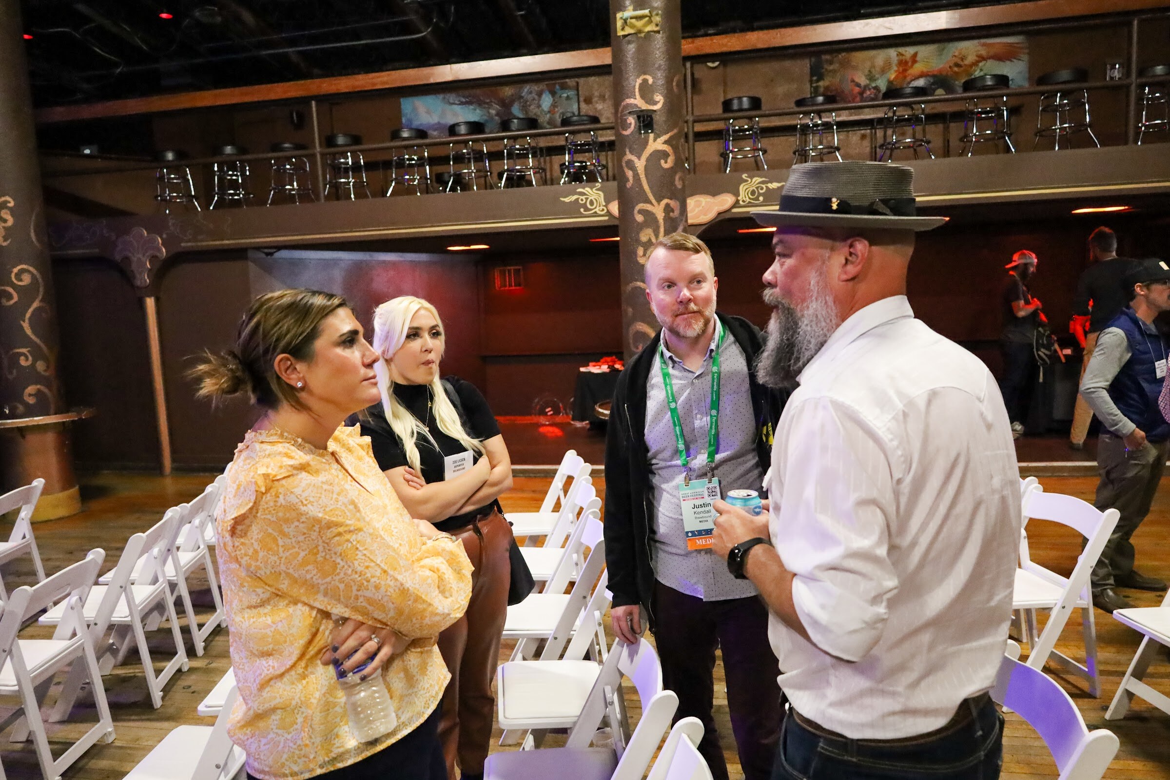 A woman in a yellow floral blouse, a woman with white-blonde hair, a man with a green conference lanyard and a bearded man in a gray hat and white shirt talk among rows of white folding chairs under the balcony.