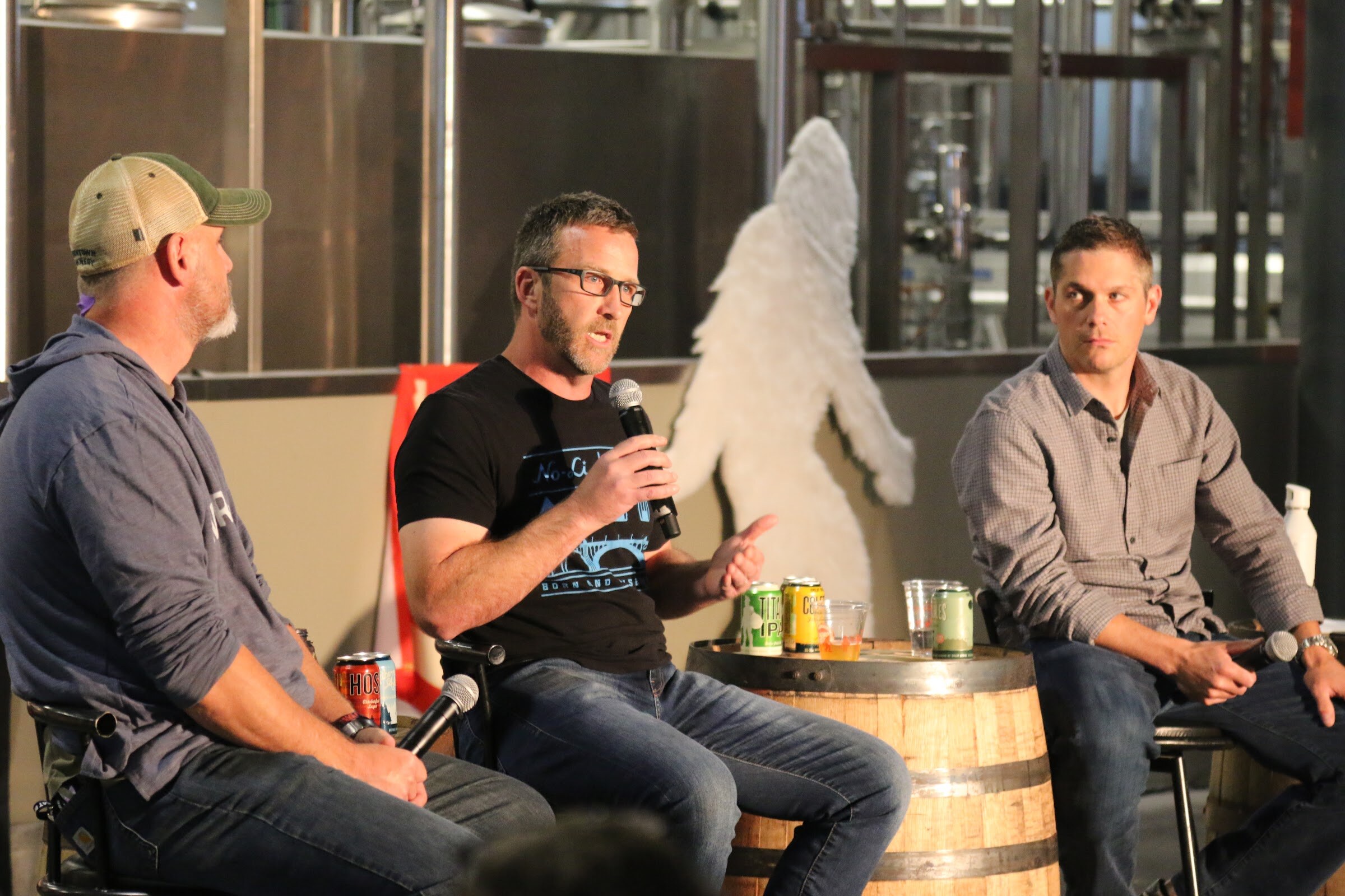 A man in black No-Li Brewhouse shirt speaks into a microphone between two other panelists, with cans on a whiskey barrel and a white Bigfoot cutout behind.