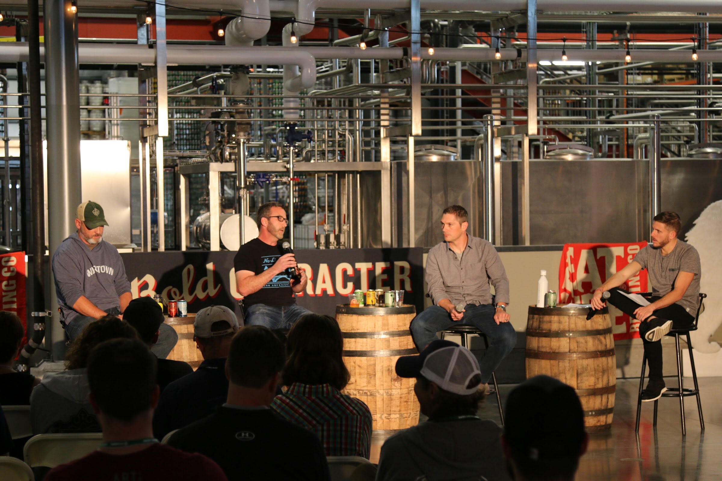 Four men on stools around two whiskey barrels with cans on them, in front of a dark Bold Character sign in a brewery full of tanks and pipes.