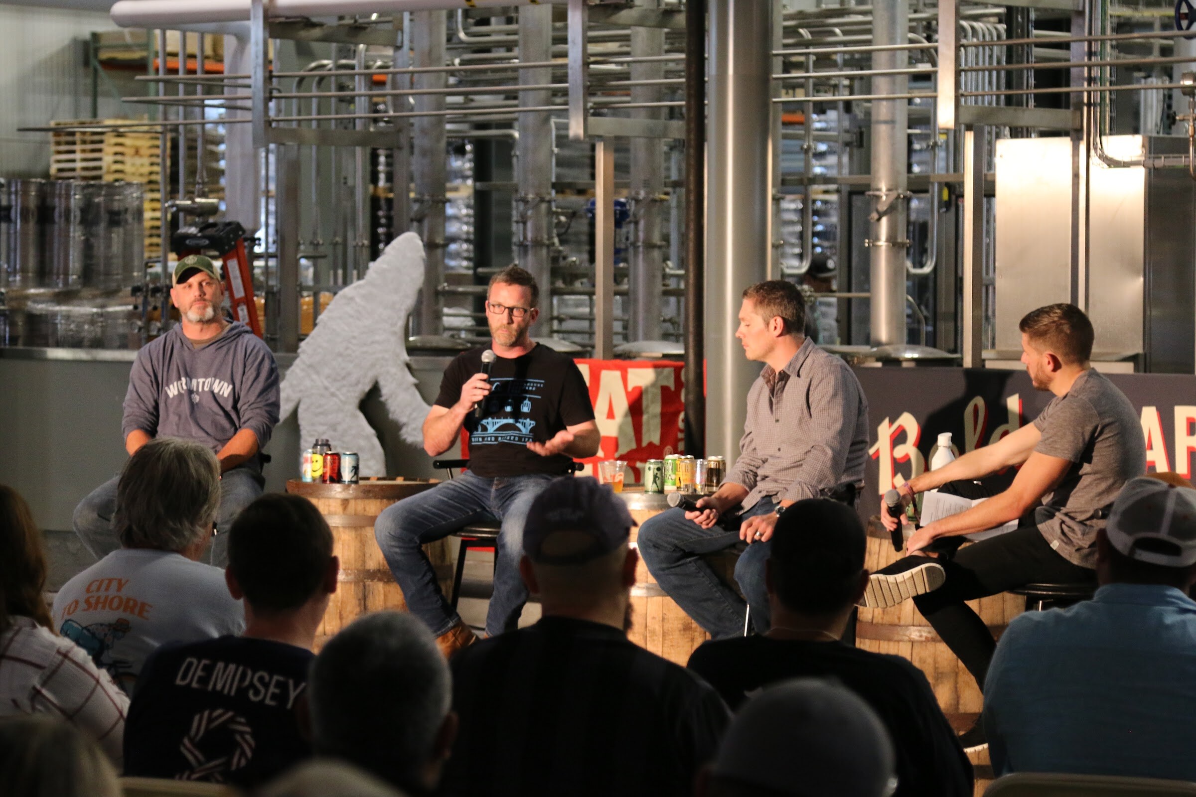Four men seated on stools around barrels on a concrete floor with brewery equipment and a Bigfoot cutout behind, and attendees seen from behind in the foreground.