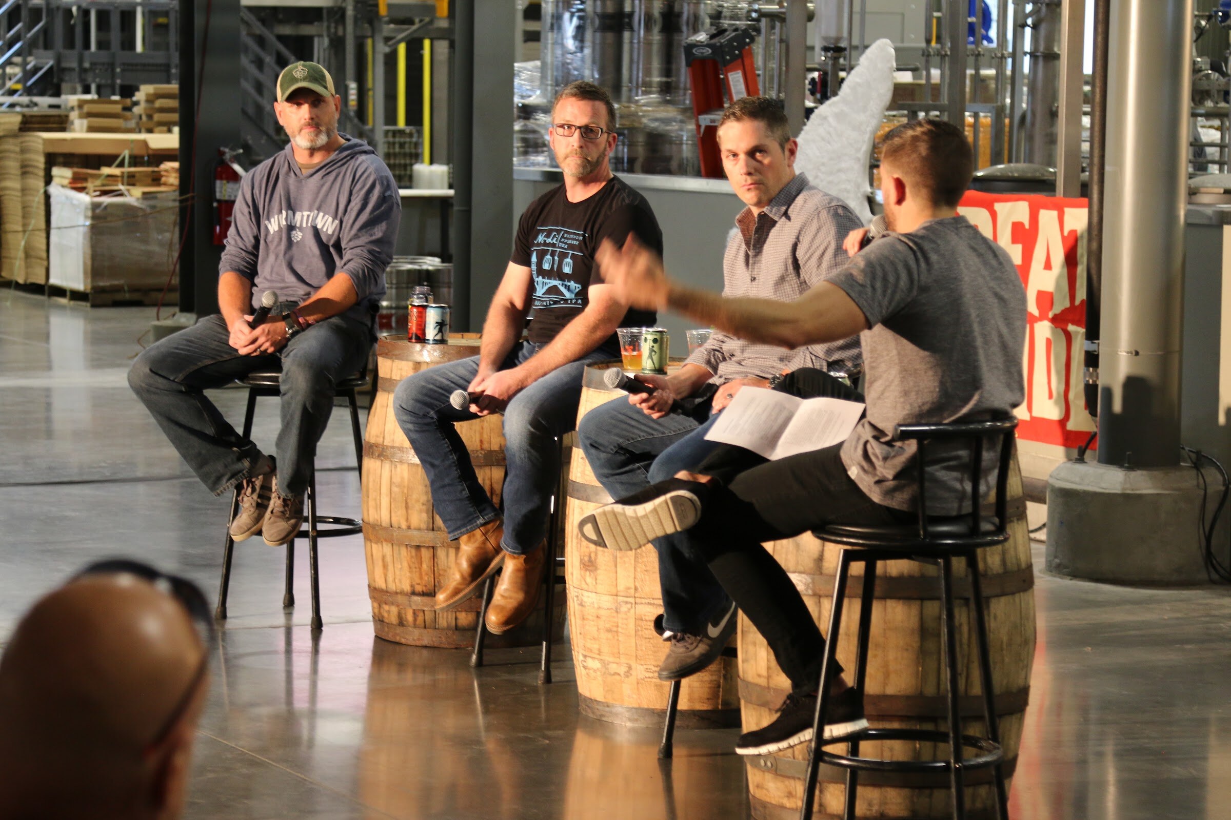 Three men on stools beside whiskey barrels listen to a fourth man who leans forward holding a microphone and a sheet of paper.