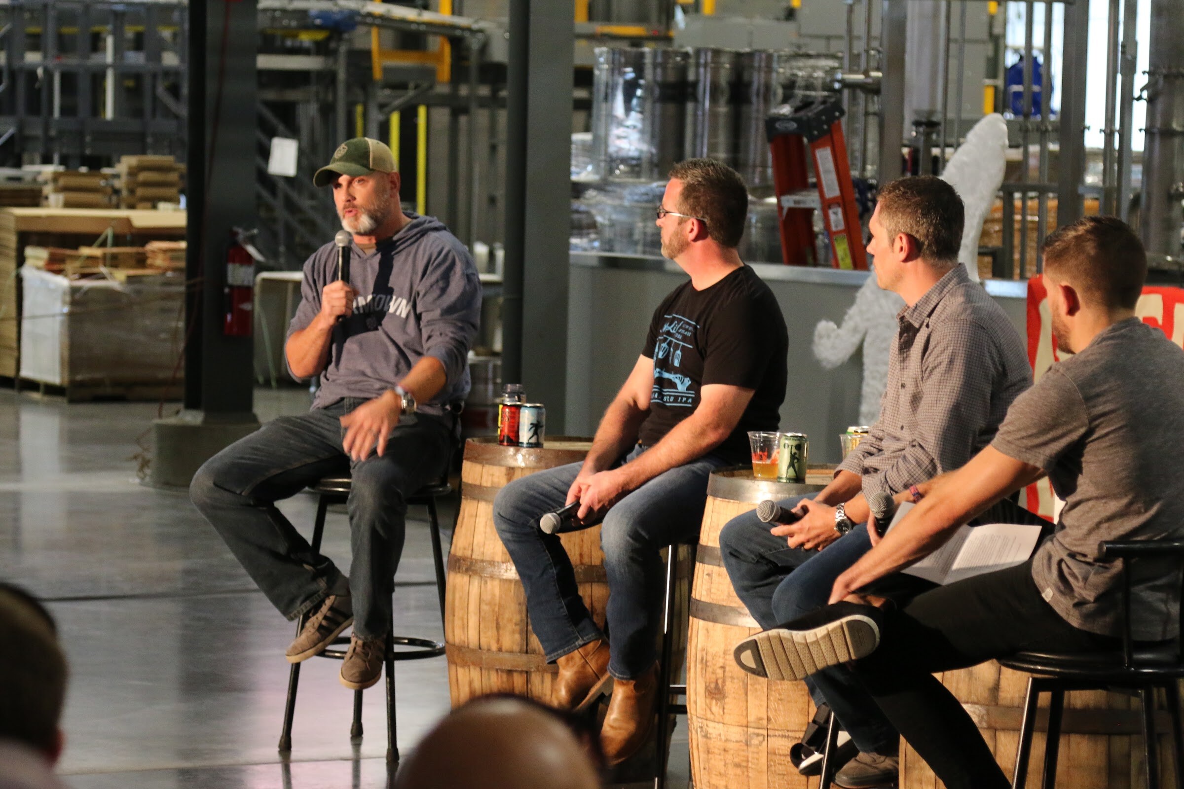 A man in a gray hooded shirt and cap speaks into a microphone on a tall stool, with three others seated beside barrels with cans on them.