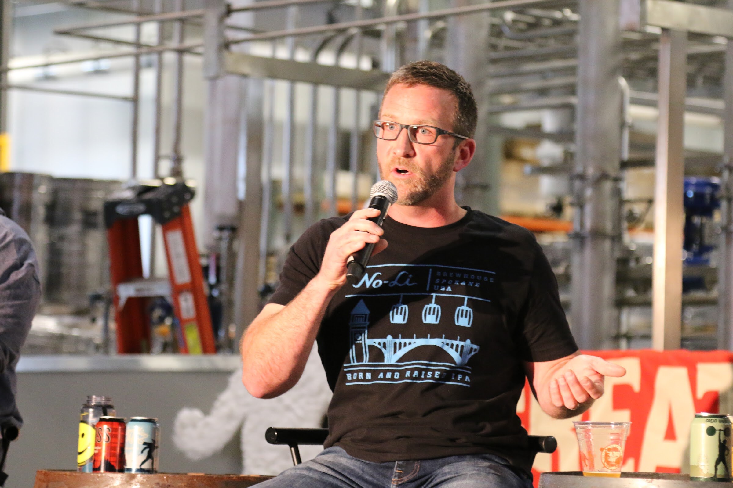 A bearded man in glasses and a black No-Li Brewhouse shirt speaks into a microphone with an open hand, with several beer cans on a barrel beside him.