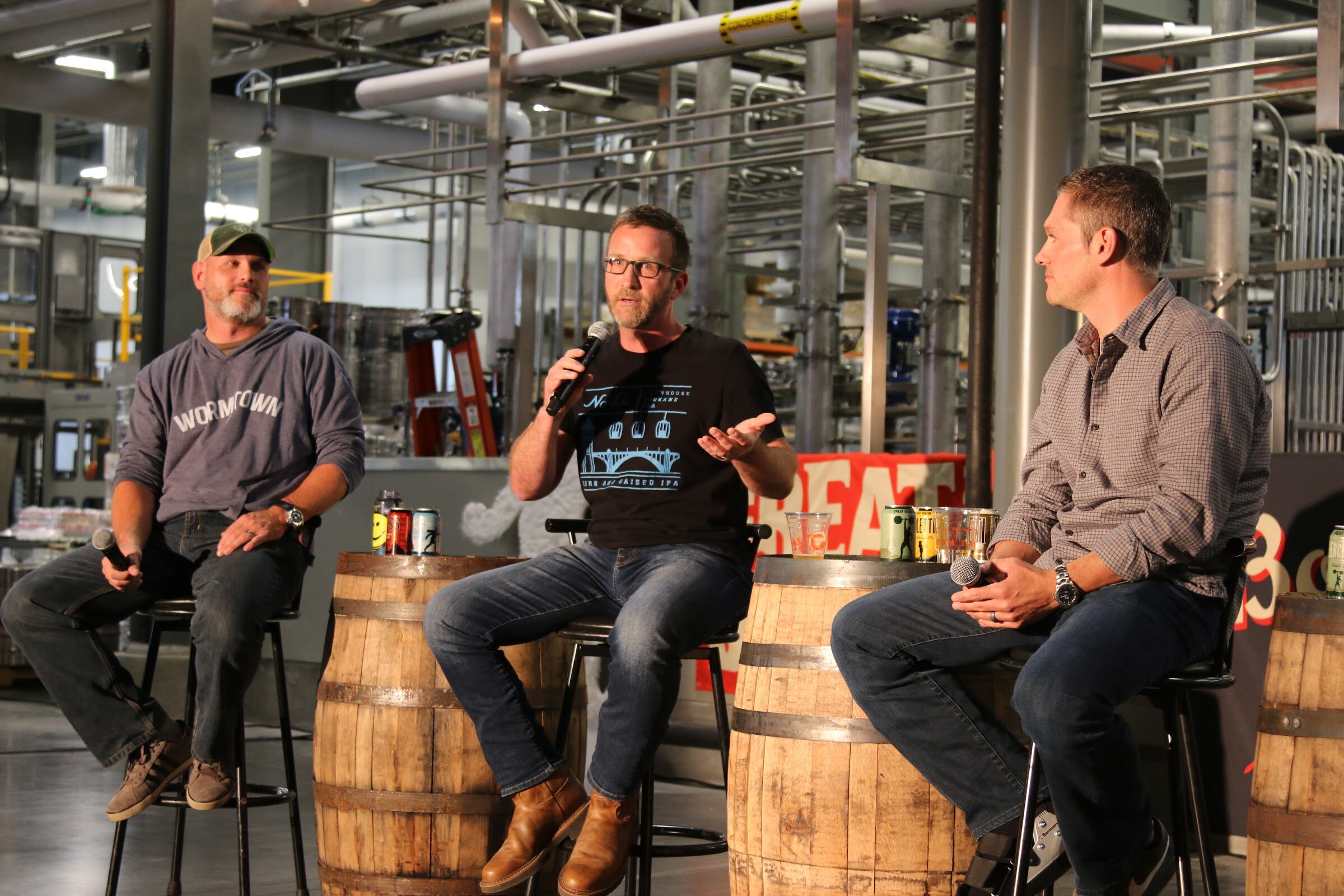Three men seated on stools beside whiskey barrels topped with cans and cups, with brewery piping and a Great Divide sign behind them.