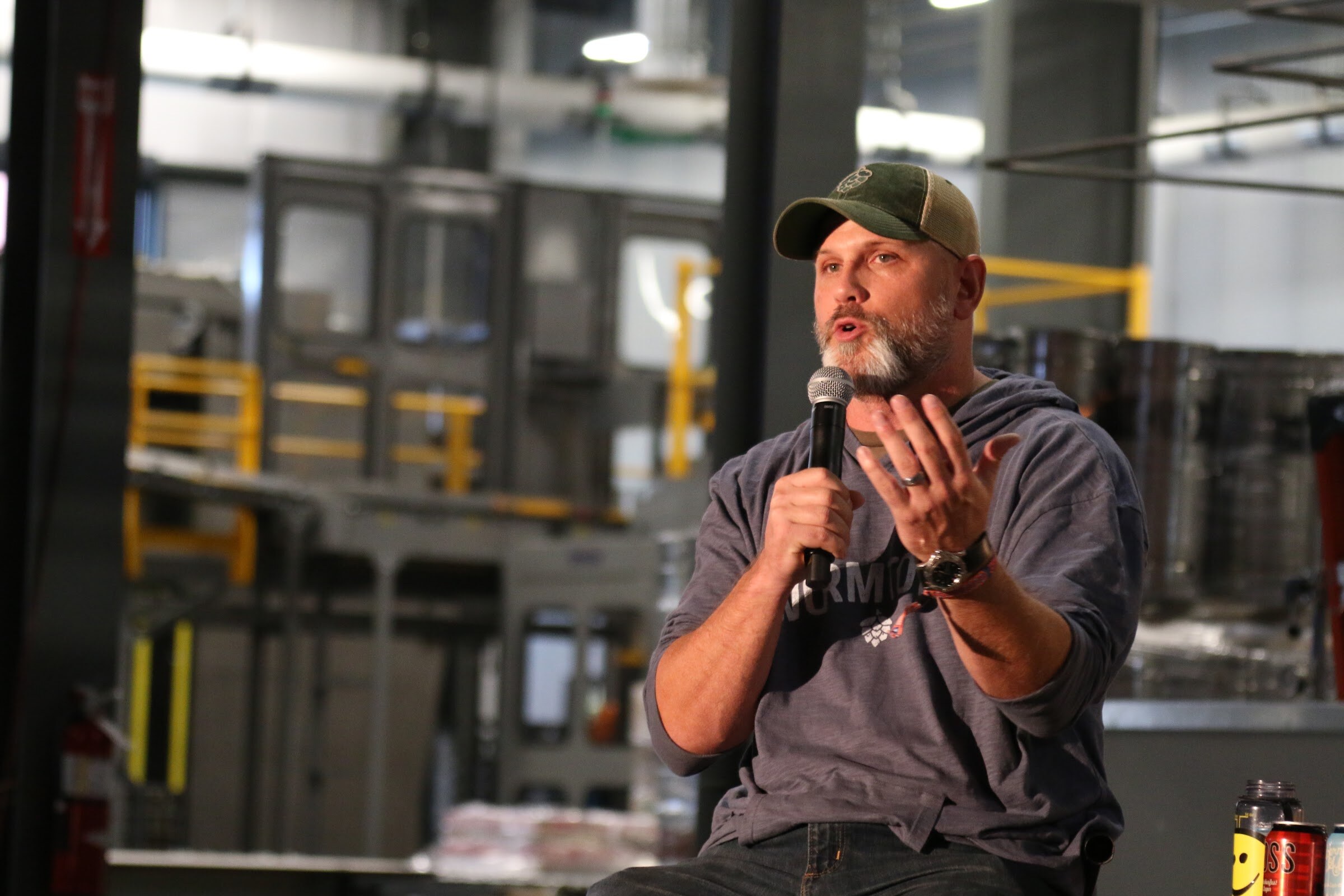 A man in a hop-logo trucker cap and gray hooded shirt speaks into a microphone with both hands raised, brewing equipment blurred behind.