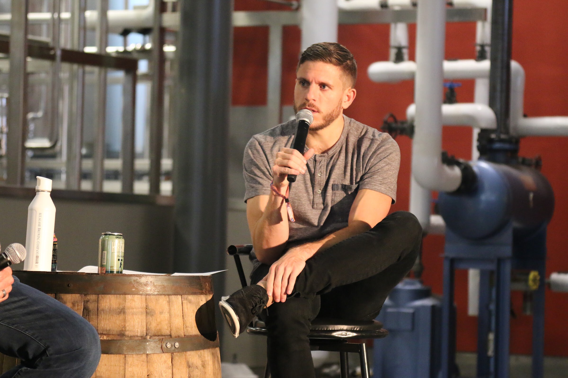 A man in a gray short-sleeved shirt sits on a stool with one leg crossed, holding a microphone beside a barrel with a can on it, against a red wall and white pipes.