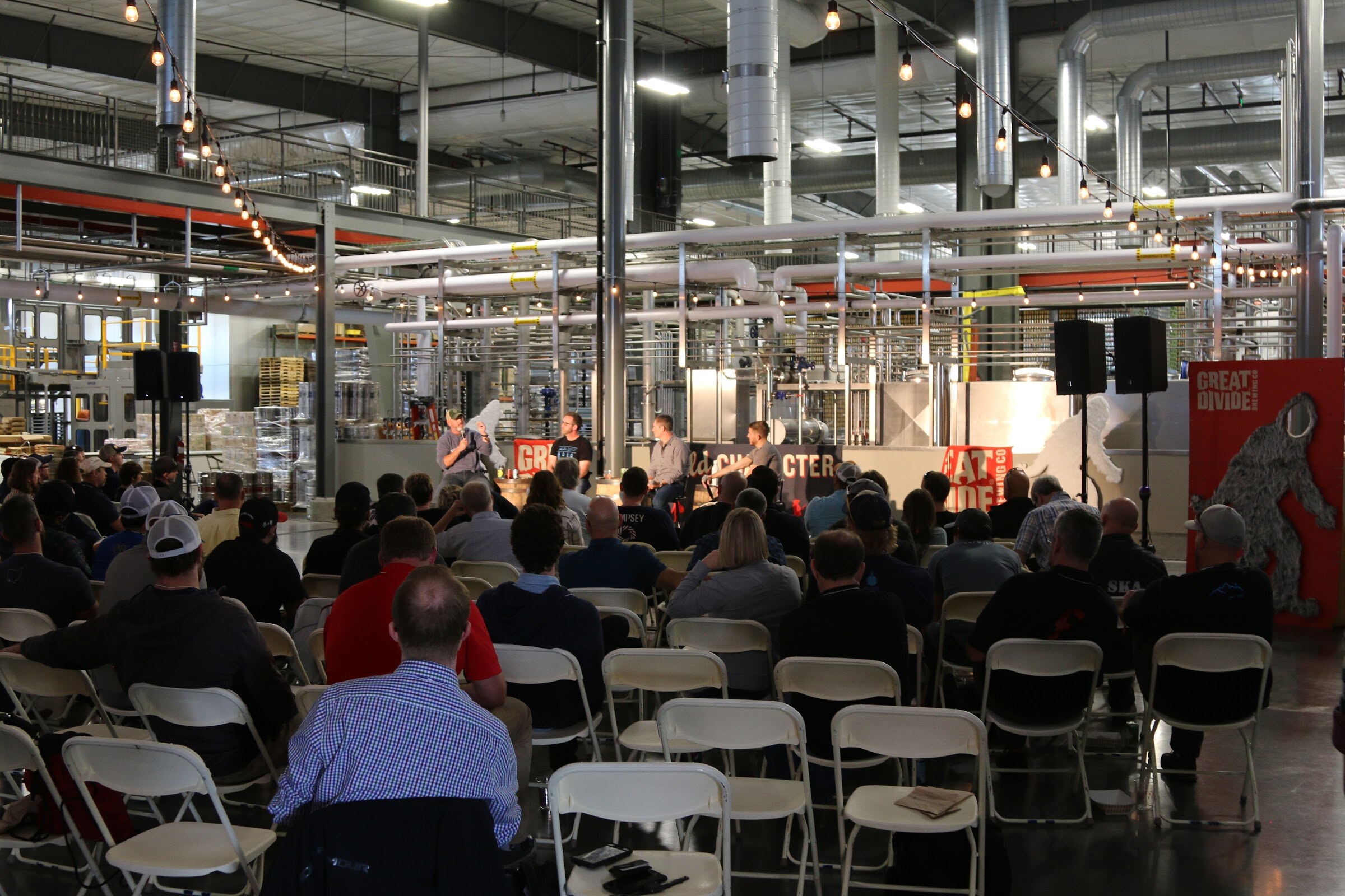 A wide view of a brewery floor with folding chairs and attendees from behind, string lights overhead and a stage with a Great Divide banner in the distance.