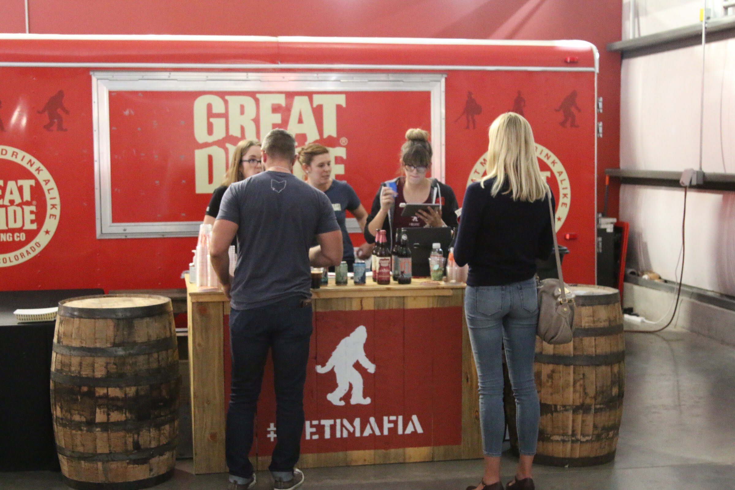 Several people stand at a wooden bar with a Bigfoot logo and Yeti Mafia lettering in front of a red Great Divide Brewing trailer.