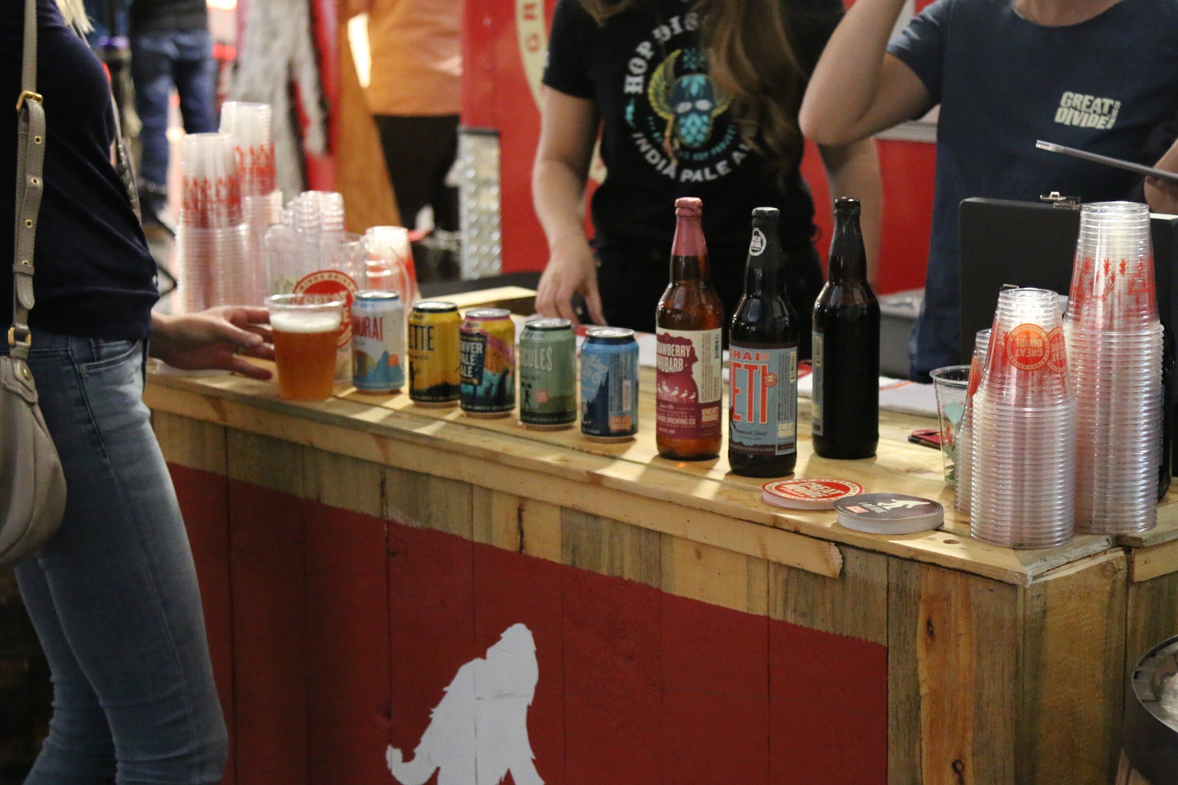 A wooden bar front-painted red with a white Bigfoot, holding a row of cans, three bottles, stacks of clear cups and a poured beer.