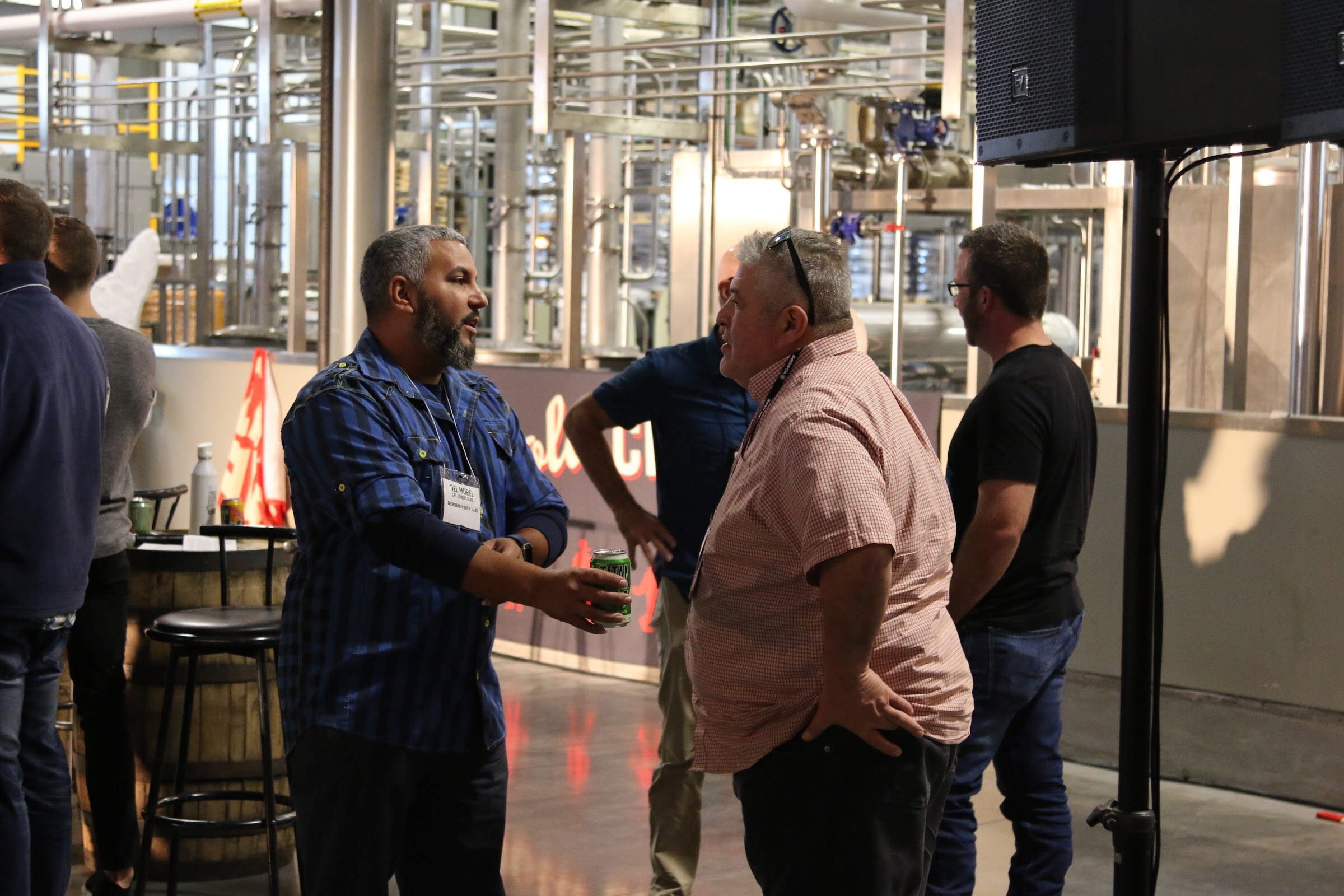 Three men in conversation in a brewery taproom area with stainless tanks, bar stools and a barrel table behind them.