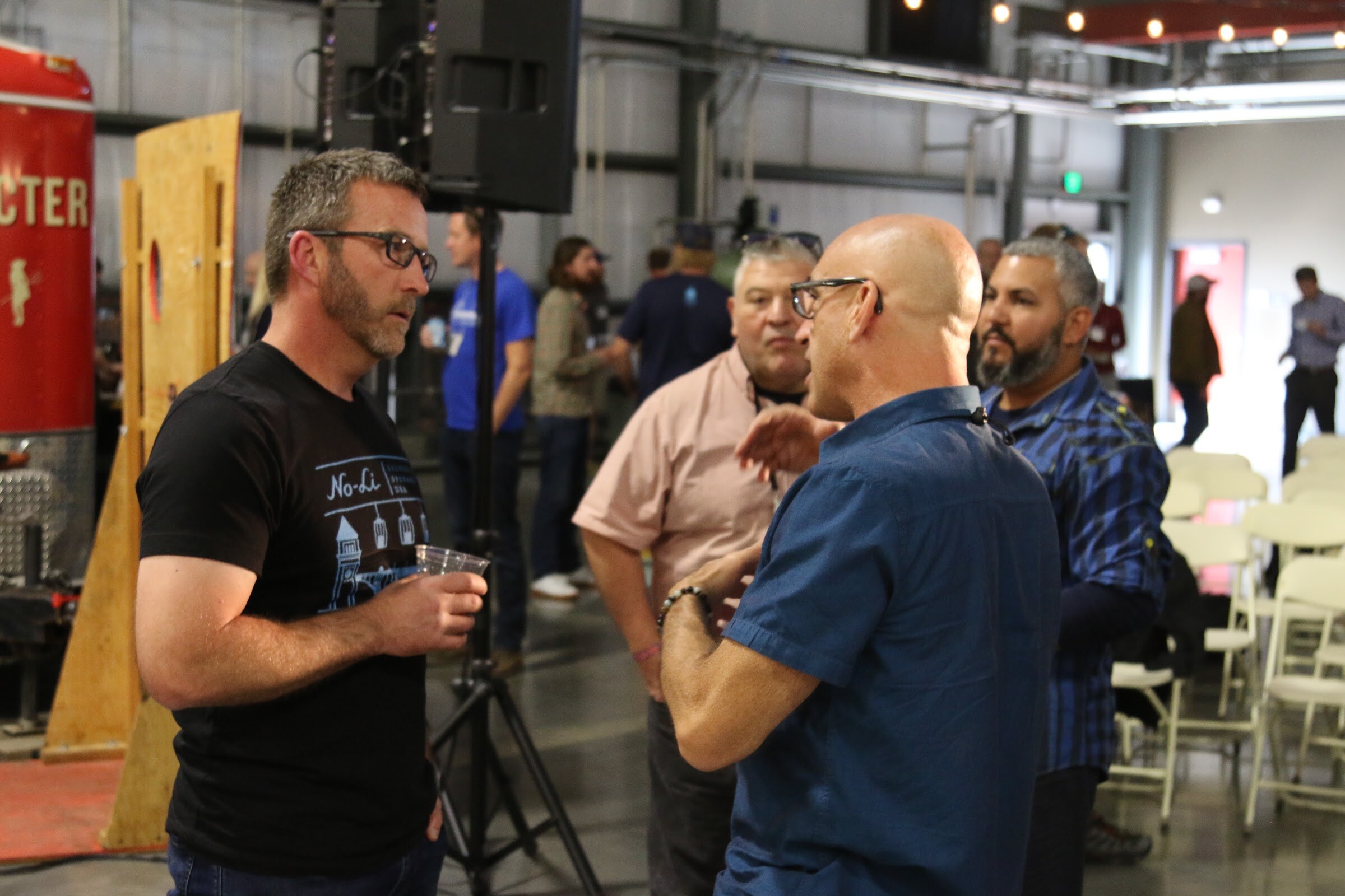 Several men in conversation, one in a No-Li Brewhouse shirt holding a clear cup, in a large brewery space with folding chairs and a red tank at the left.