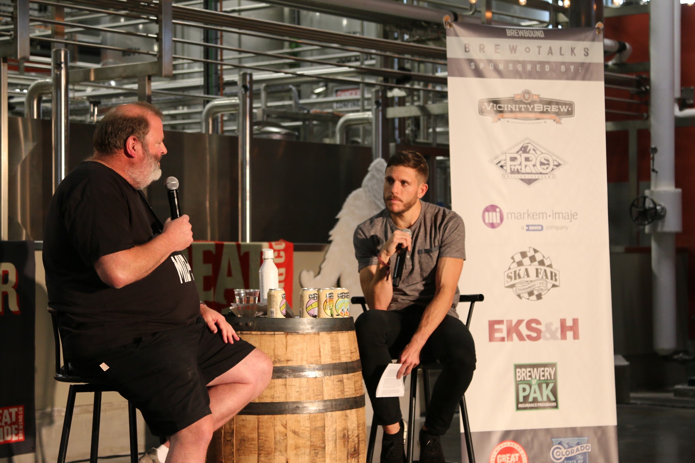 Two men on stools by a whiskey barrel with cans on it, with a tall white Brew Talks banner listing VicinityBrew, Ska Fab and Brewery PAK.