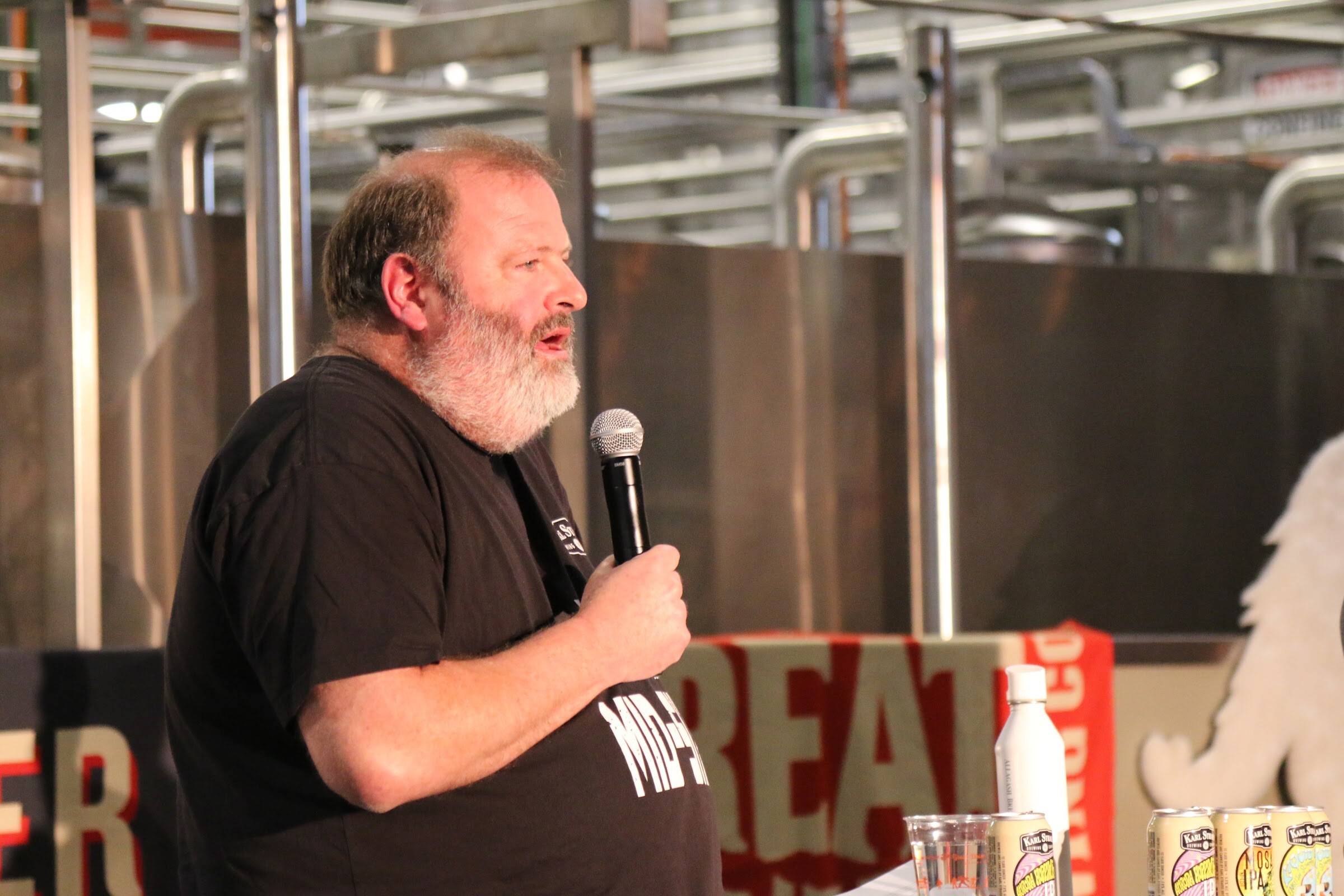 A close view of a bearded man in a black Mid-Size t-shirt speaking into a microphone, with a barrel with cans and a Great Divide sign behind him.