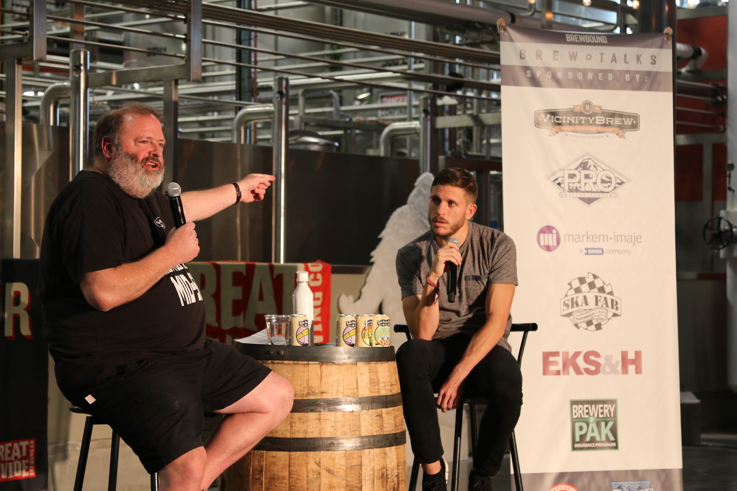 A bearded man in a black shirt points across the room with a microphone in his other hand, while a younger man in gray holds a microphone beside a white Brew Talks banner.