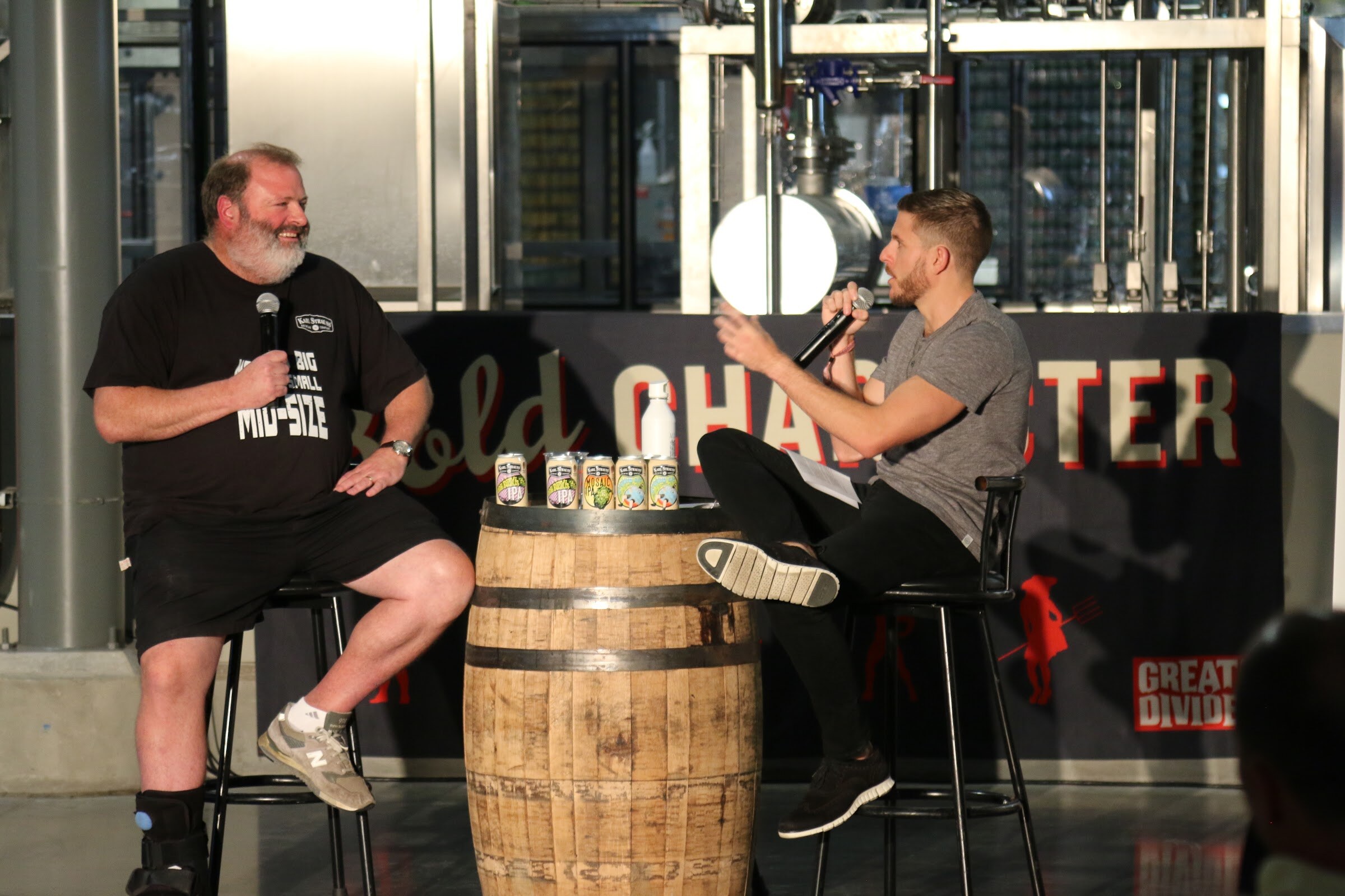A smiling bearded man with a microphone sits on a stool beside a whiskey barrel while a man in a gray shirt leans forward with a microphone.