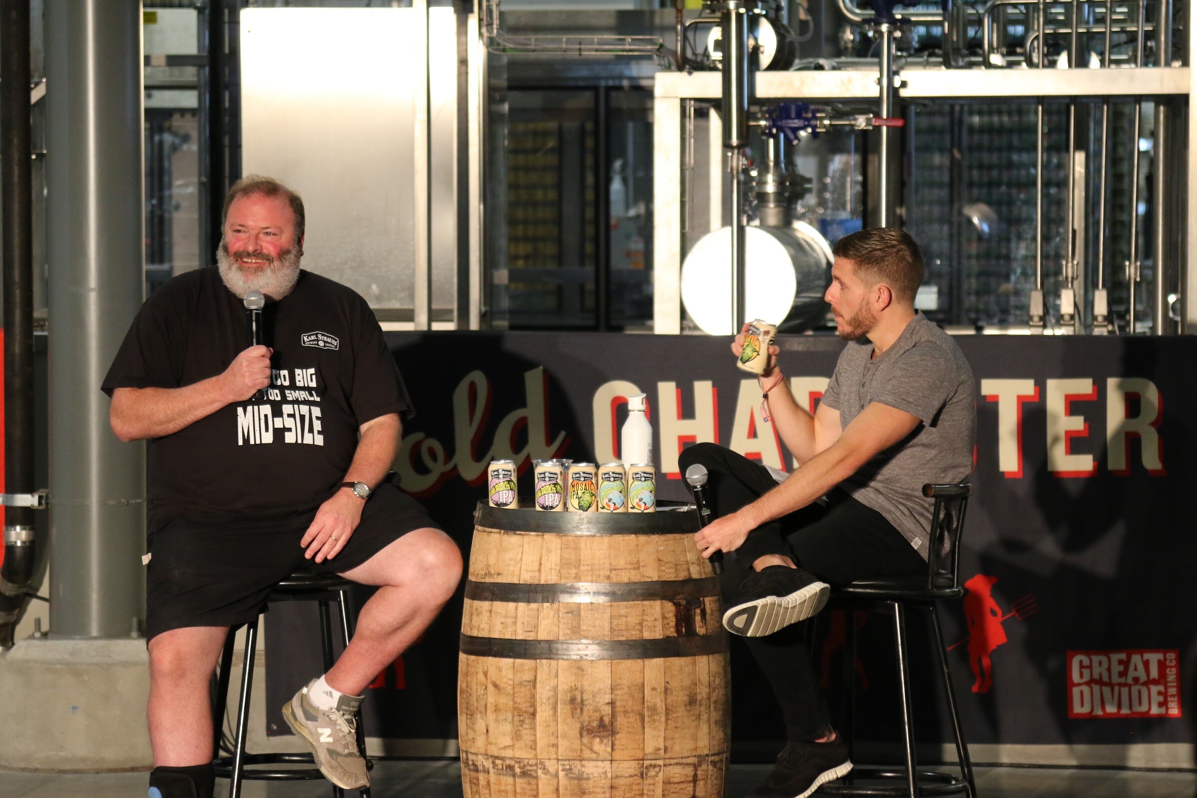 A smiling bearded man in a black Mid-Size shirt sits by a whiskey barrel while a man in gray holds up a can, with a Bold Character sign behind.