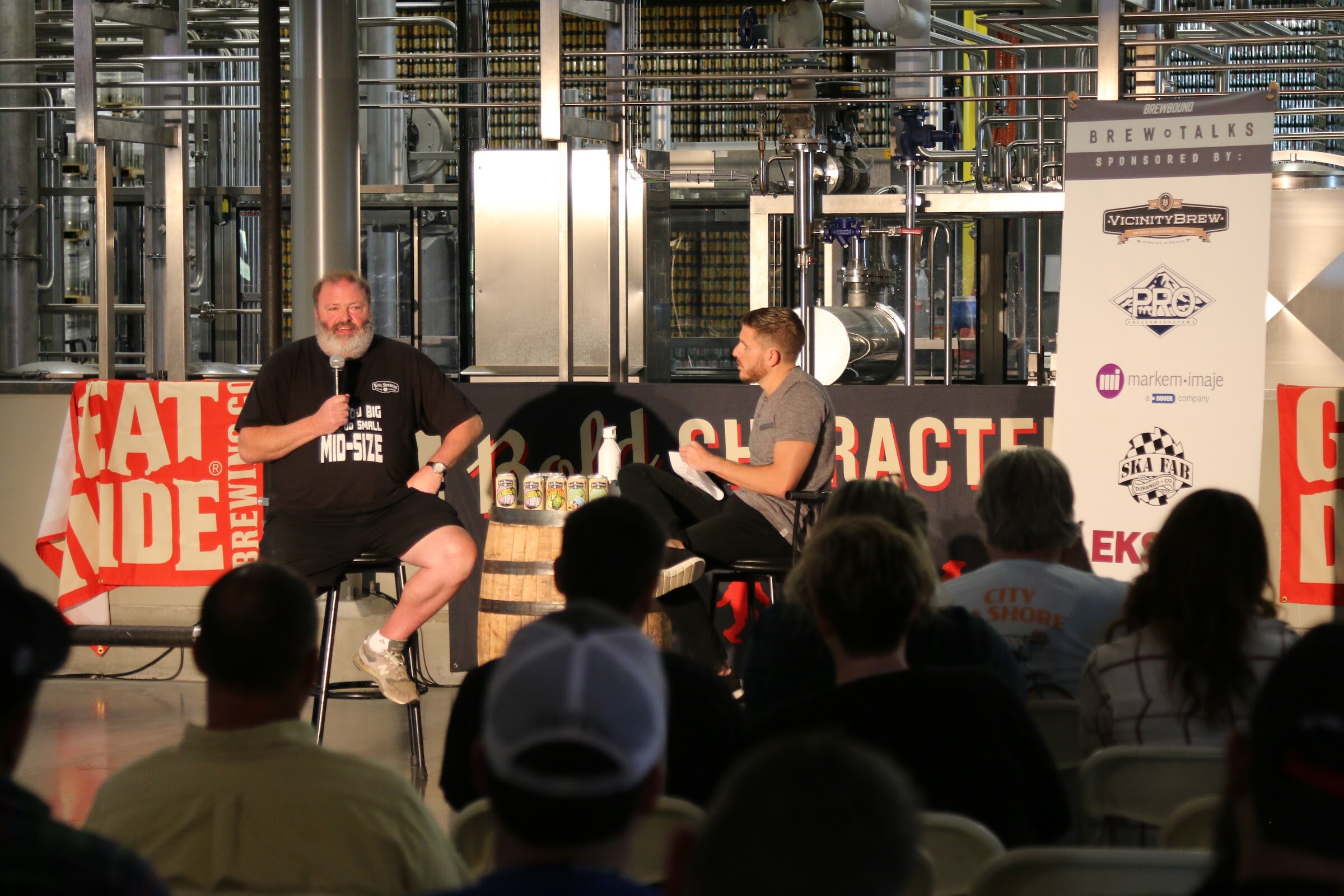 Attendee heads and caps in the foreground facing a stage where a bearded man and a moderator sit by a whiskey barrel in front of a Brew Talks banner.