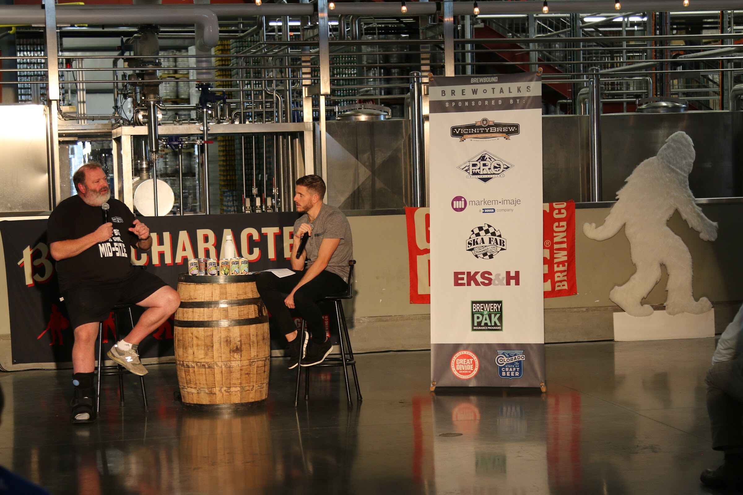Two men seated on stools beside a whiskey barrel in a brewery, with a white Brew Talks banner and a large white Bigfoot cutout beside them.