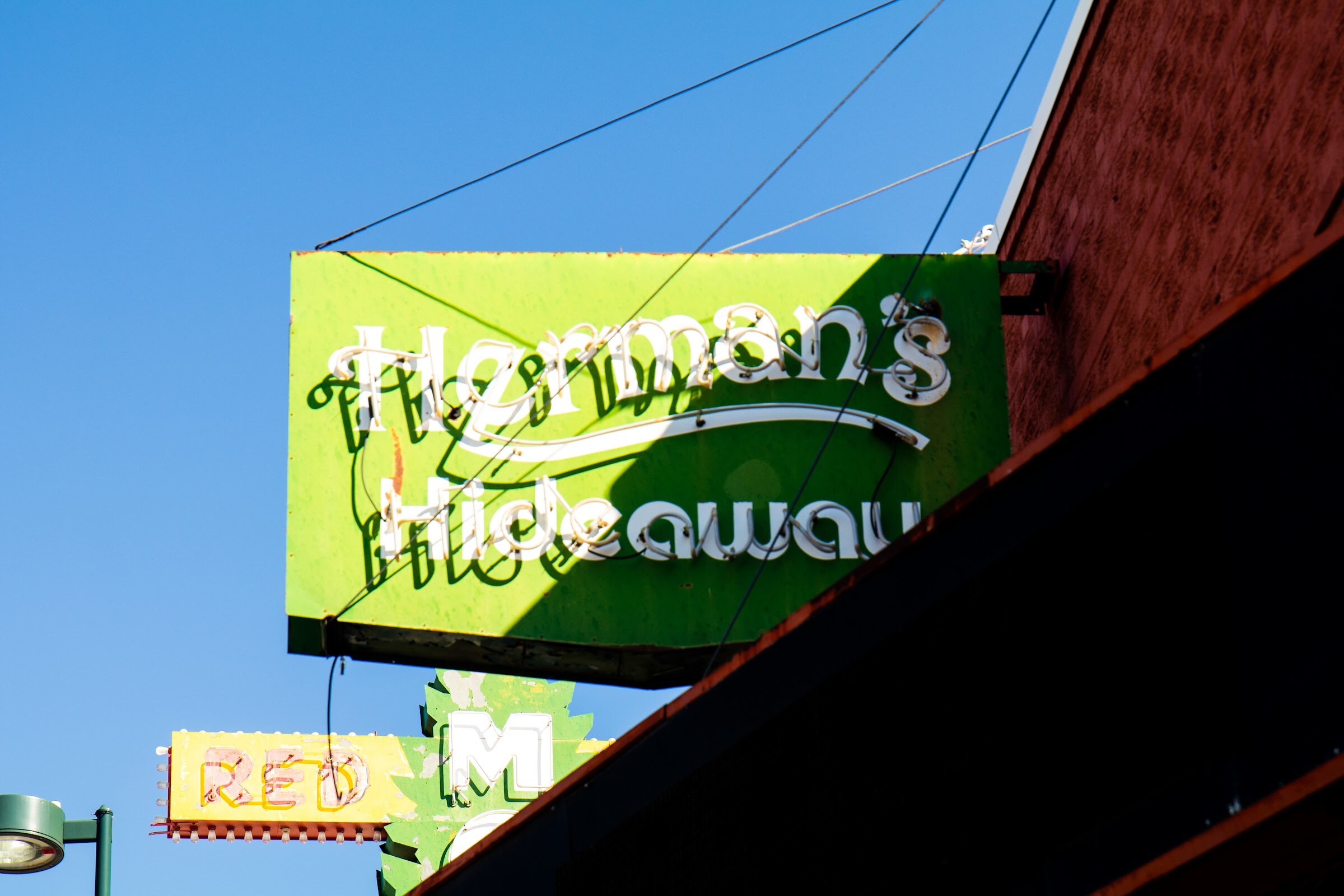 A green neon sign reading Herman's Hideaway against a blue sky, with a smaller Red M sign below it.