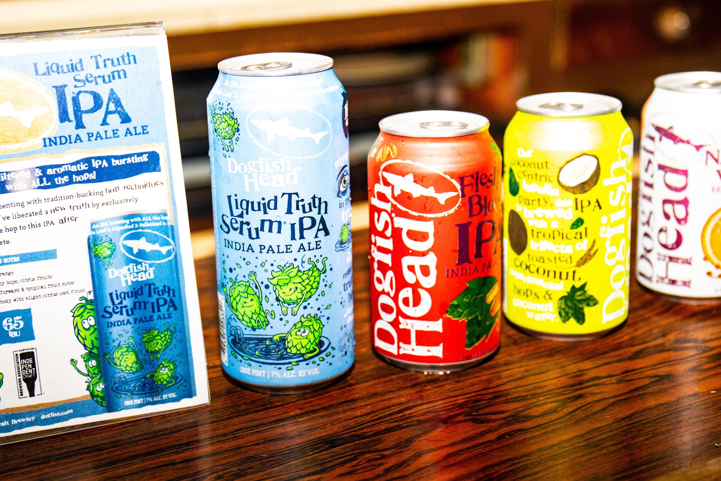 Four Dogfish Head cans in blue, red, yellow and white on a polished wood bar next to a printed Liquid Truth Serum IPA card.