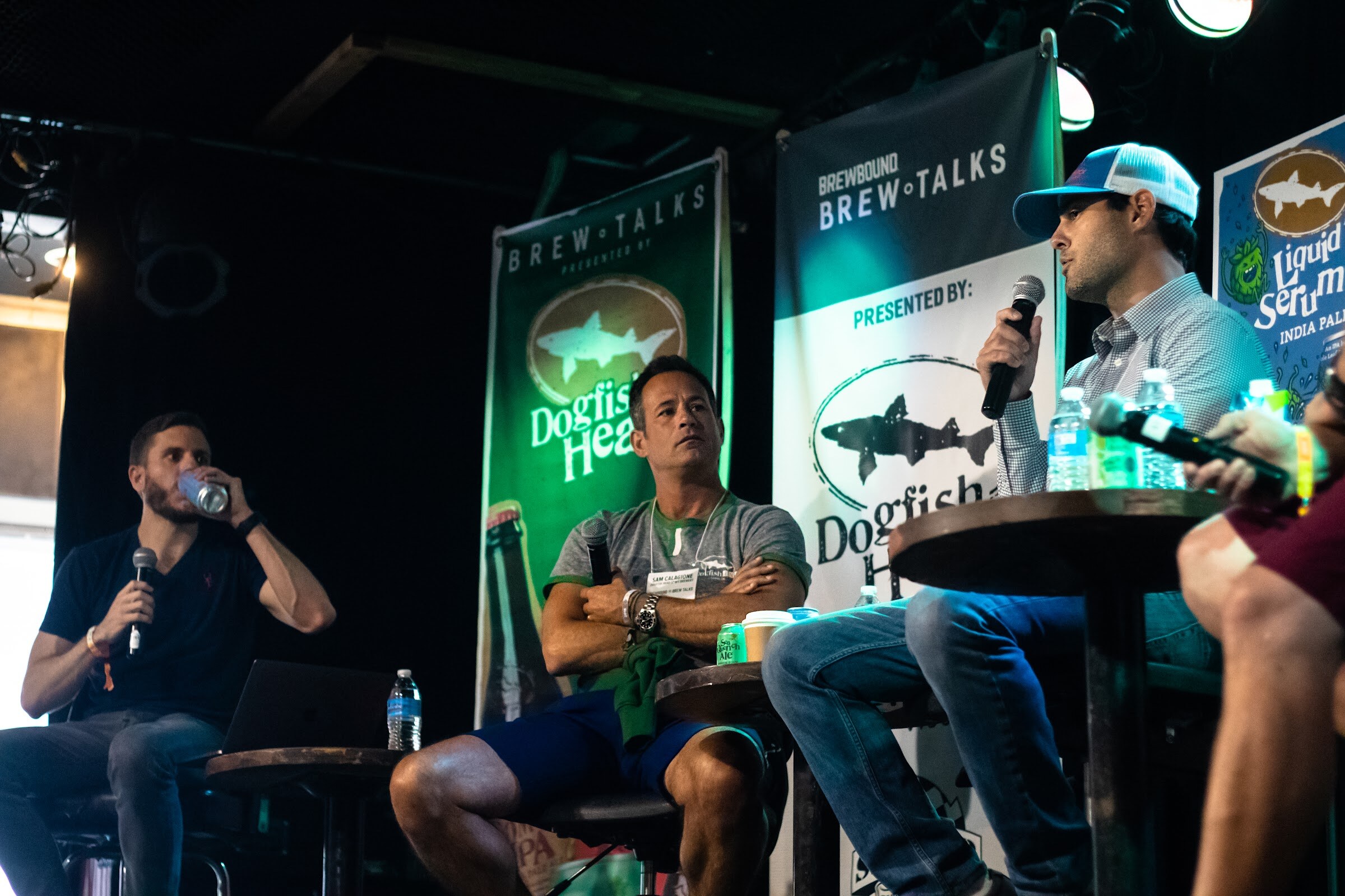 Three men on stools on a dark stage with a Dogfish Head banner, a Brewbound Brew Talks banner and a Liquid Truth Serum IPA poster; one drinks from a can, one sits with arms crossed, one speaks into a microphone.