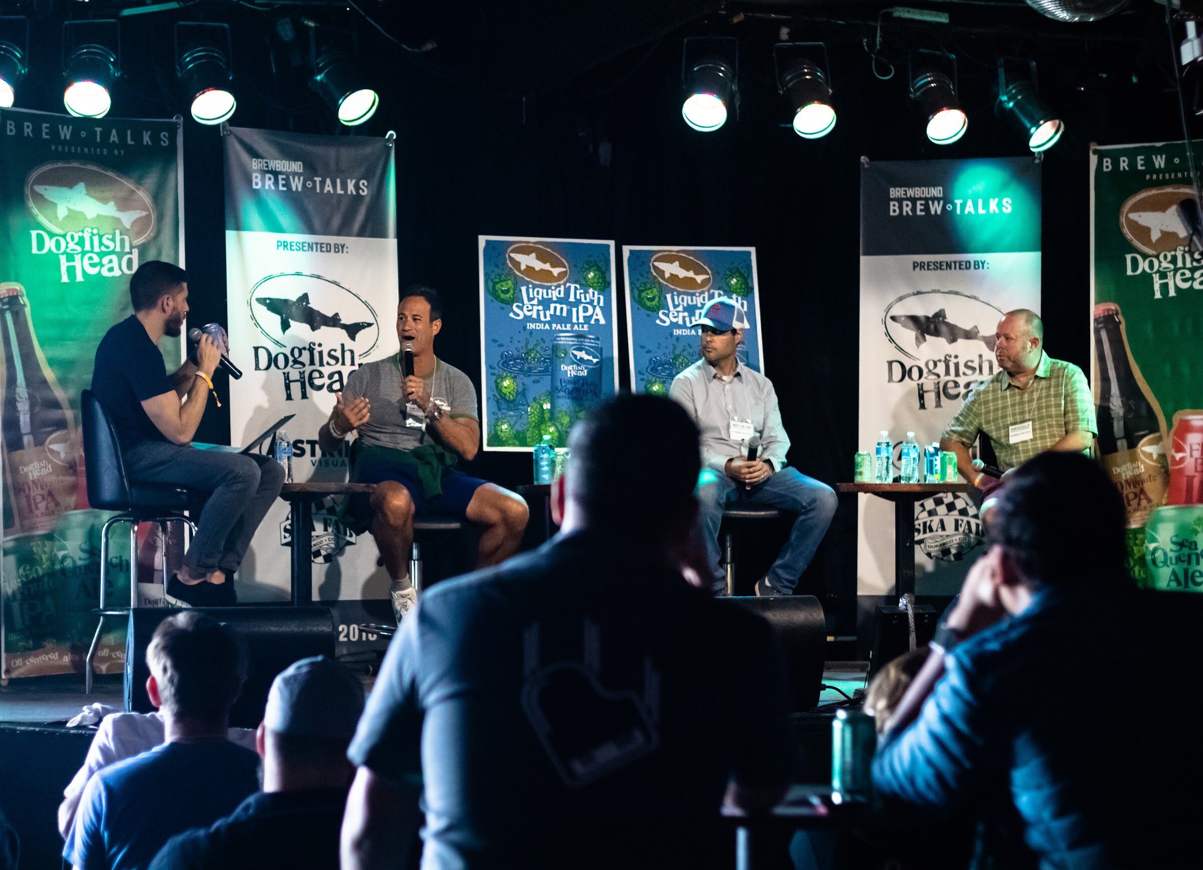 A moderator with a microphone, two panelists and a third panelist at a small table on a stage with Brew Talks and Dogfish Head banners and Liquid Truth Serum IPA posters; the backs of audience heads are in the foreground.