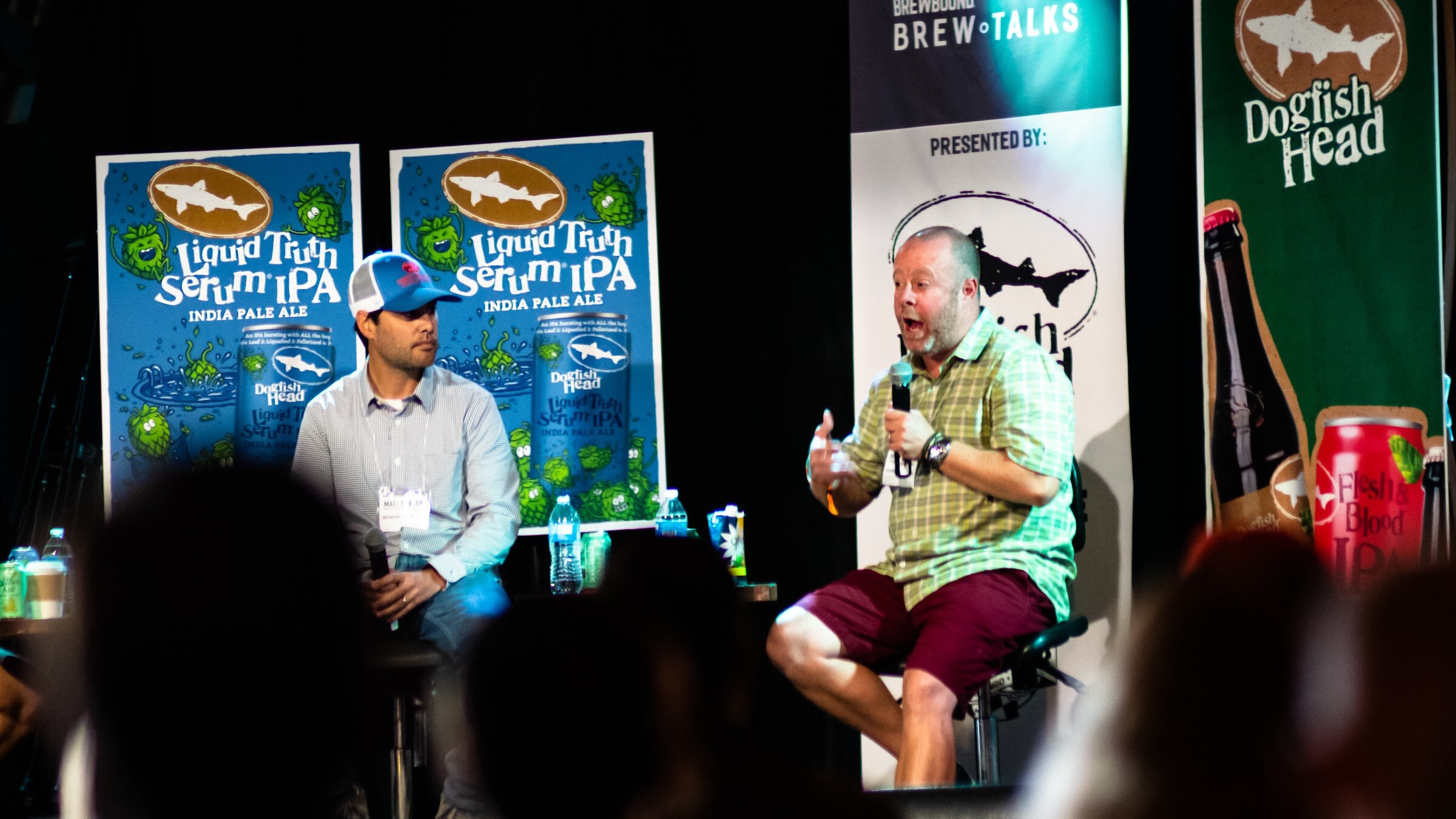 A man in a green check shirt and burgundy shorts speaks into a microphone on a stool next to a man in a blue cap, with Liquid Truth Serum IPA posters and a Dogfish Head banner behind them.