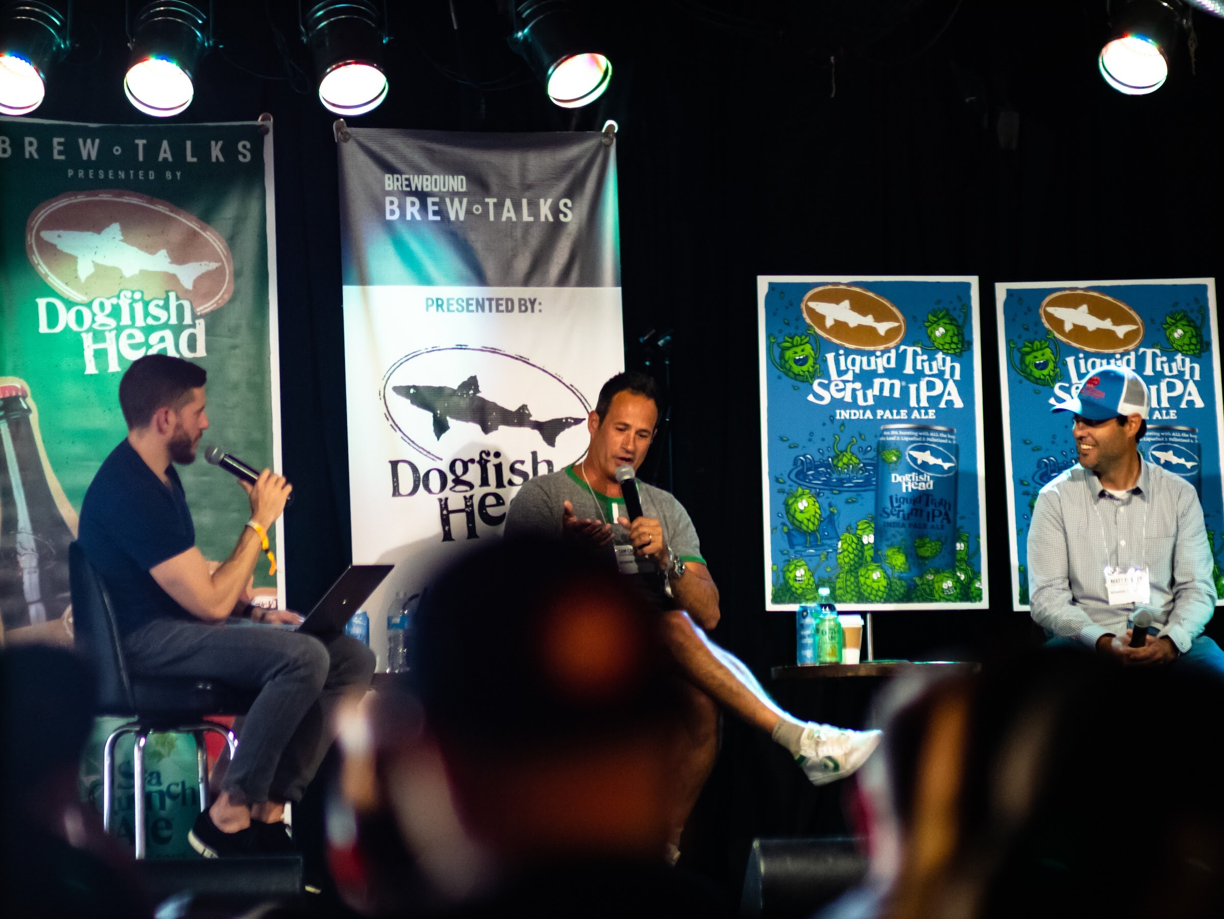 Three men on a stage with Dogfish Head and Brew Talks banners and Liquid Truth Serum IPA posters; the one in the middle speaks into a microphone.