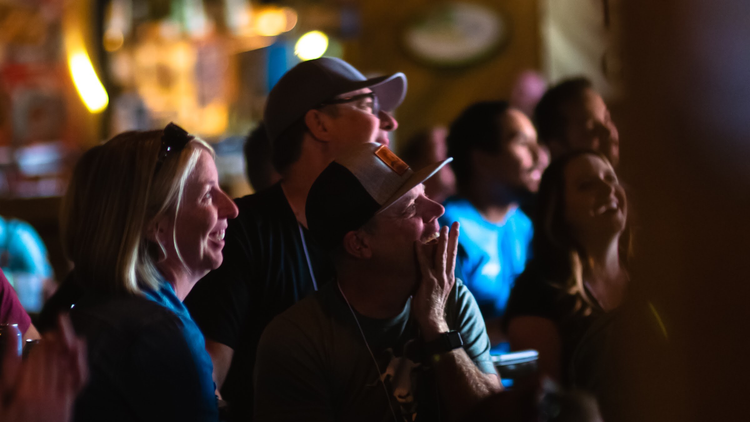 A dim, close crop of a crowd of attendees, several in caps, laughing and smiling as they look toward the stage.