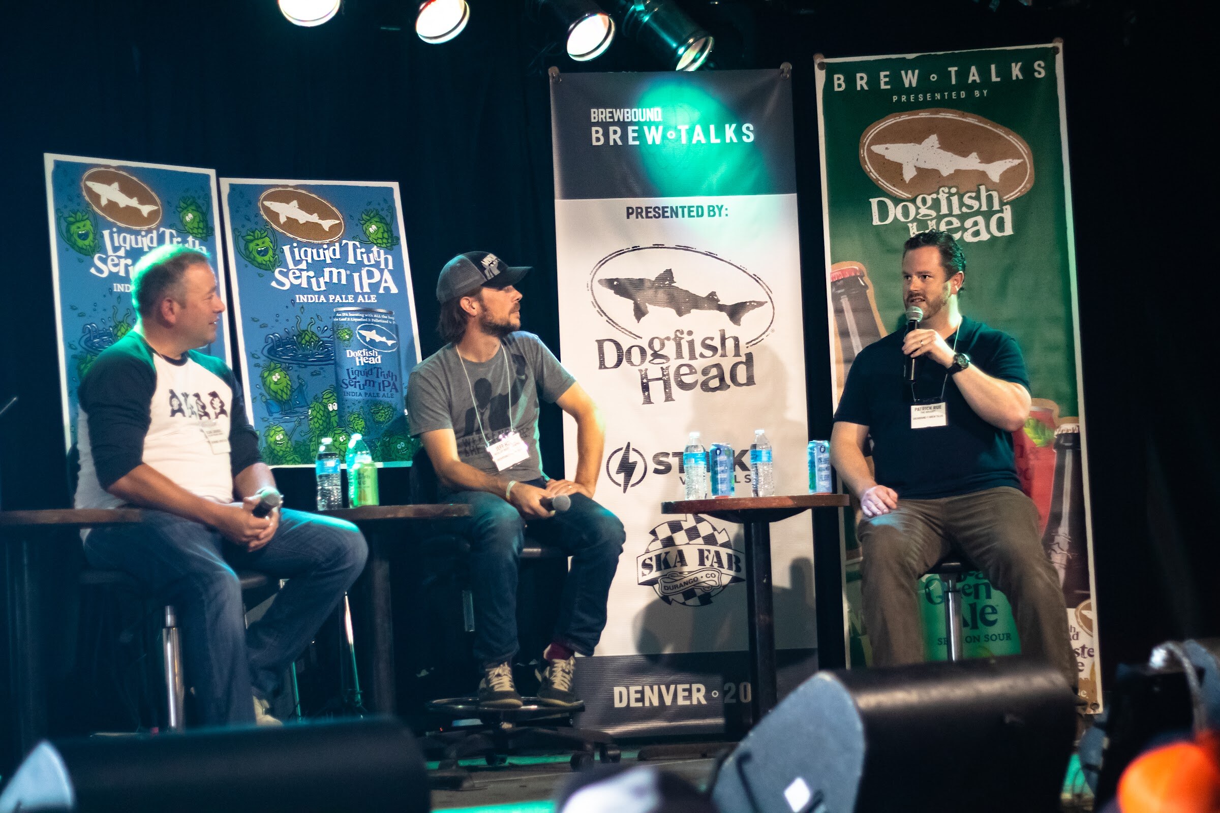 Two men and a third man speaking into a microphone sit on stools on a stage with a Brewbound Brew Talks banner reading Denver, a green Dogfish Head banner and Liquid Truth Serum IPA posters.