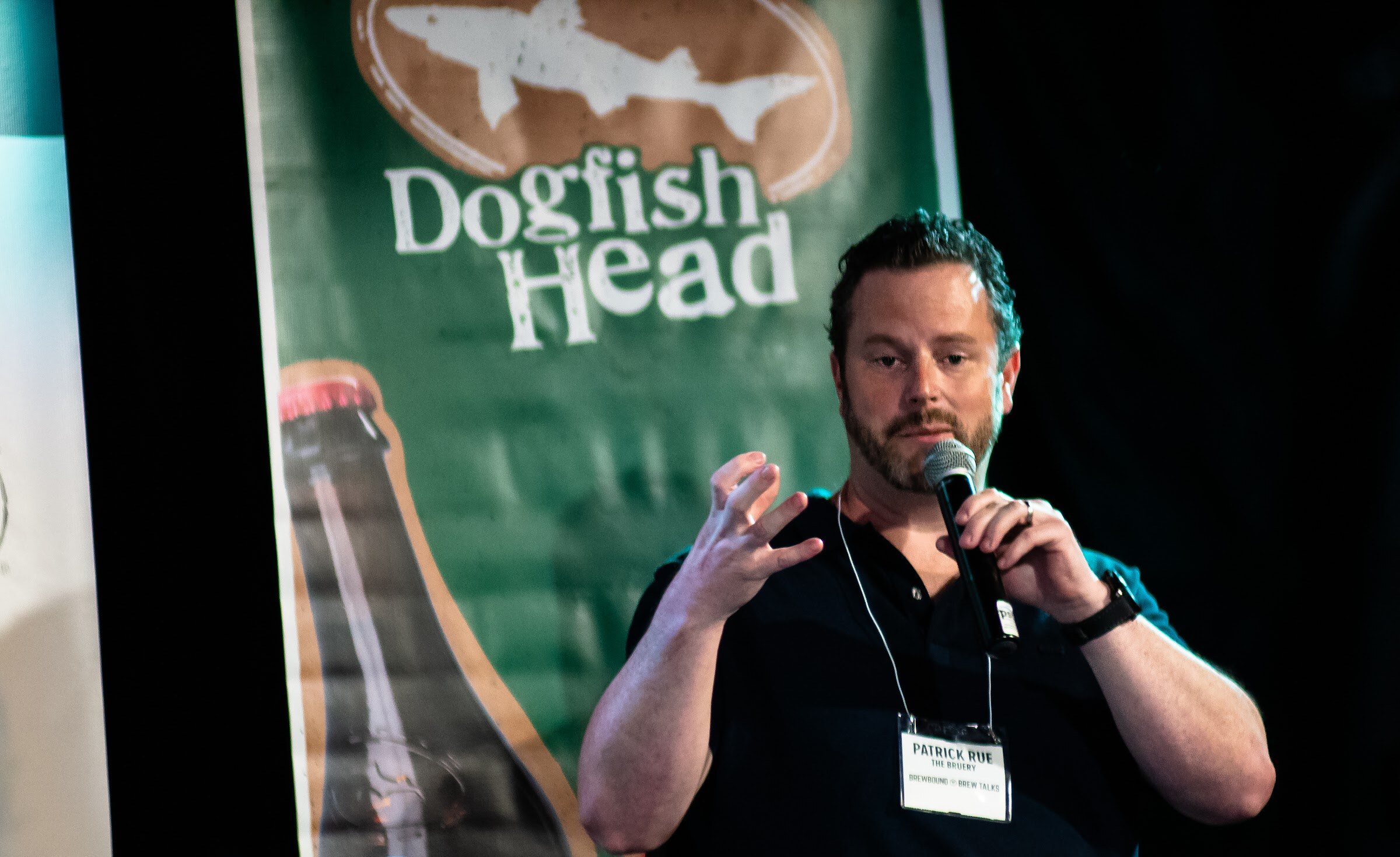 A bearded man in a dark polo shirt with a lanyard holds a microphone and gestures in front of a green Dogfish Head banner.