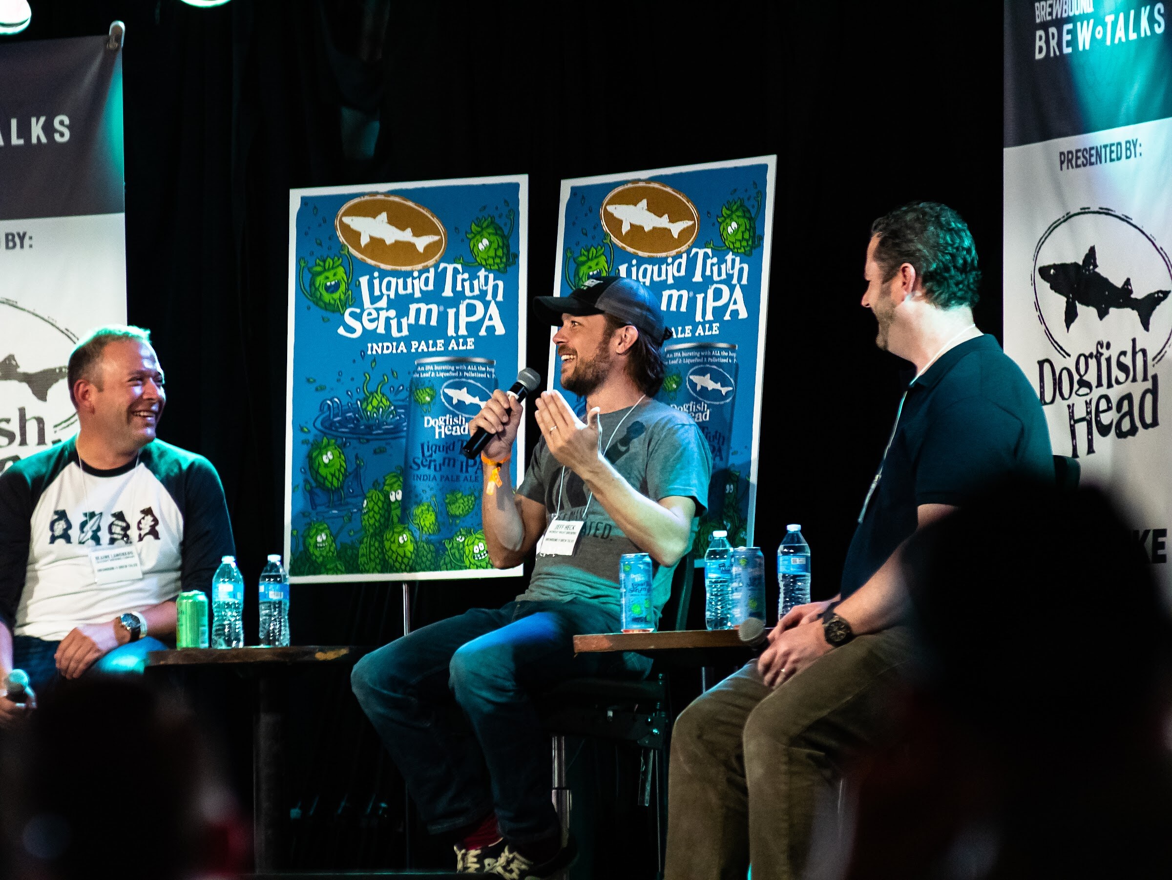 A man in a cap and gray T-shirt speaks into a microphone in front of Liquid Truth Serum IPA posters, with a man in a baseball shirt laughing at left and a dark-haired man listening at right.