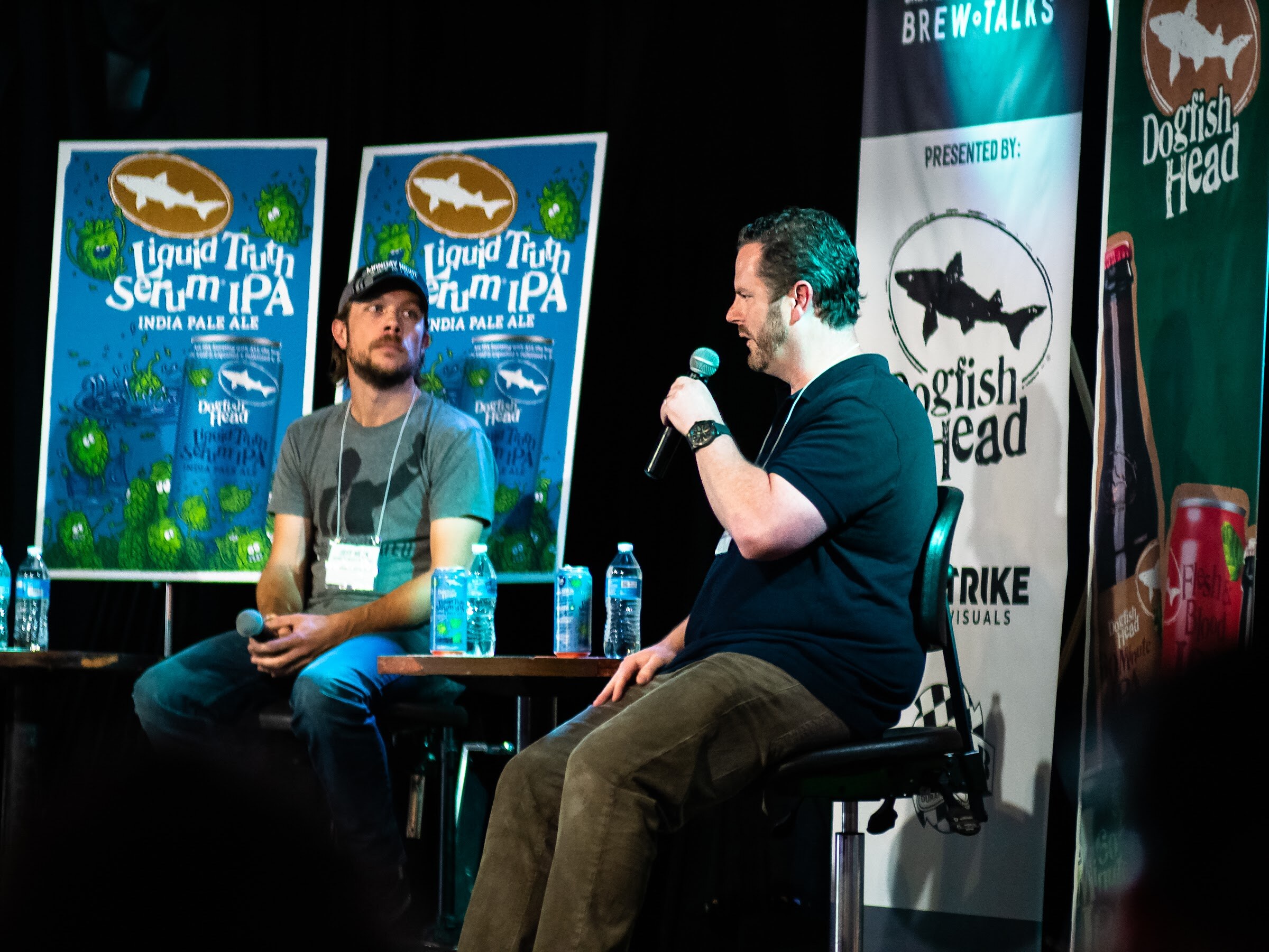 Two men on stage; one in profile speaks into a microphone, the other in a gray T-shirt and cap listens, with Liquid Truth Serum IPA posters and Dogfish Head banners behind them.