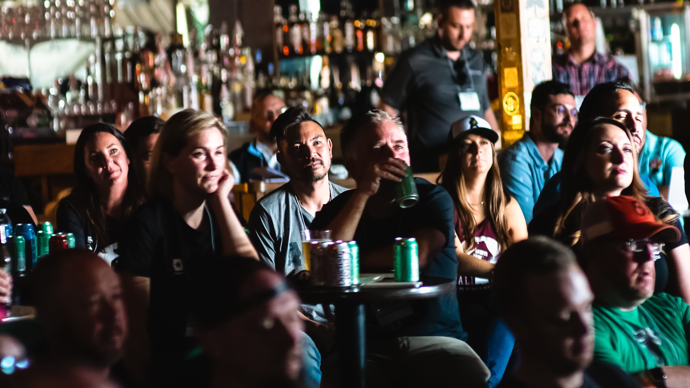 A dim, crowded bar with seated and standing attendees, some resting a chin on a hand, with green beer cans on small tables and a lit bar behind them.